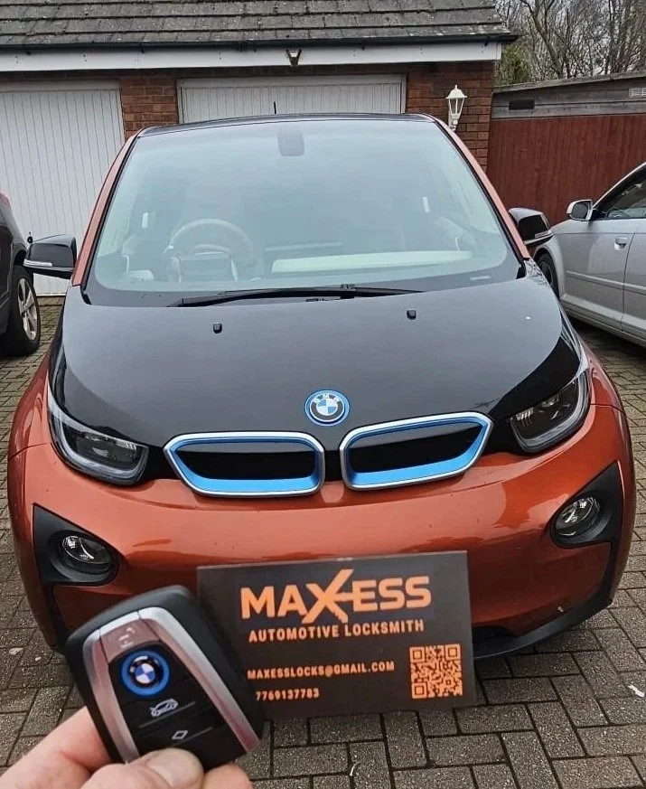 Front view of a BMW i3 electric car with a black hood and blue accents on the grille, parked on a driveway with a person holding a BMW key fob and a business card for Maxess Automotive Locksmith in front of the car.