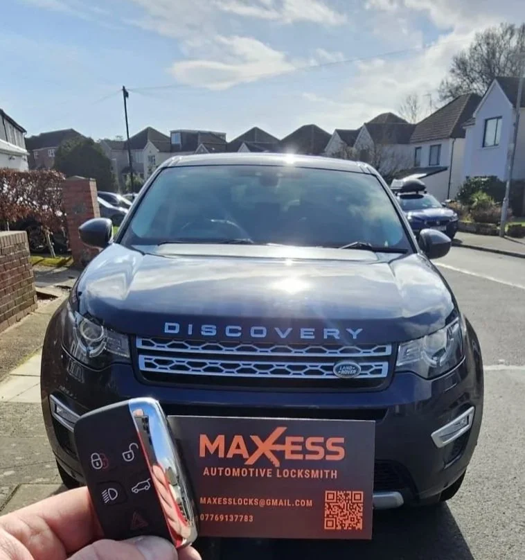 A person holding a car key with a discovery vehicle in the background. The vehicle is parked outside in a residential neighborhood and has a business card from Max Ess Automotive Locksmith placed on its front bumper.