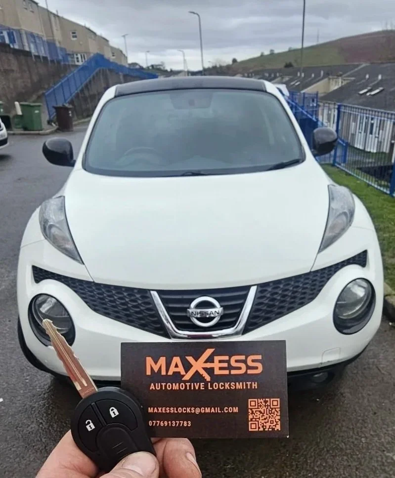 Front view of a white Nissan Juke parked outdoors on a cloudy day with a person holding a car key and a business card for Maxess Automotive Locksmith.