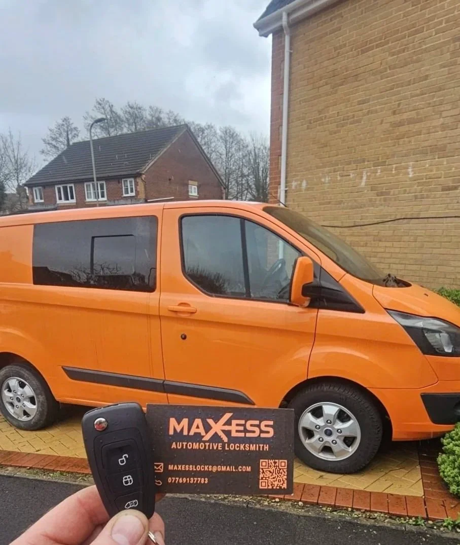 A person holds a car key fob with multiple buttons in front of an orange van parked beside a business card on the ground. The business card reads 'MAXESS AUTOMOTIVE LOCKSMITH' with contact information. In the background, there are residential buildin