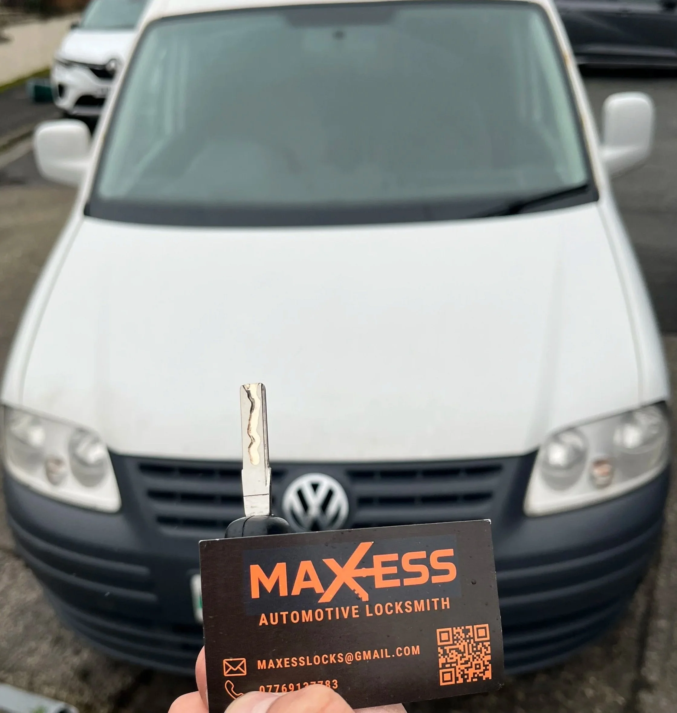 A person holding a Maxess Automotive Locksmith business card and a car key in front of a white Volkswagen vehicle.