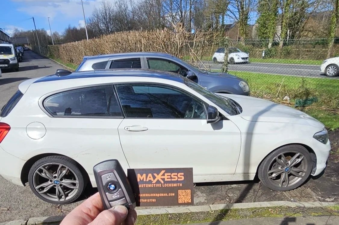 A white BMW coupe parked outdoors with a black and orange business card for Maxess Automotive Locksmith placed on the ground in front of it. The person holding a BMW key fob is visible in the foreground. Several other cars are parked or driving in th