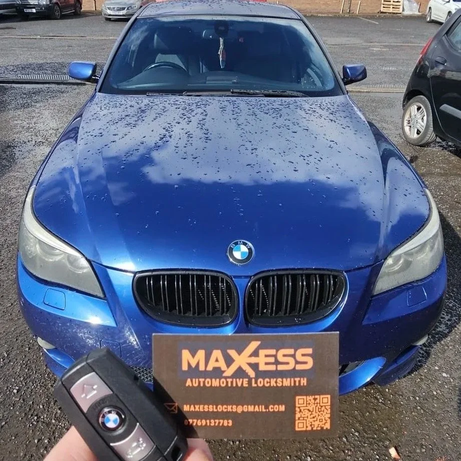 Blue BMW car parked outdoors on a gravel lot with water droplets on the hood, a sign from Maxess Automotive Locksmith, and a person holding a BMW key fob in the foreground.