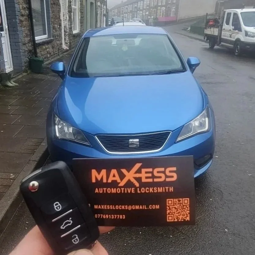 A blue car parked on a misty street with brick houses in the background. Someone is holding a car key and business card for Maxess Automotive Locksmith in front of the car.
