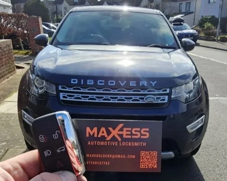A person holding a car key with a discovery vehicle in the background. The vehicle is parked outside in a residential neighborhood and has a business card from Max Ess Automotive Locksmith placed on its front bumper.