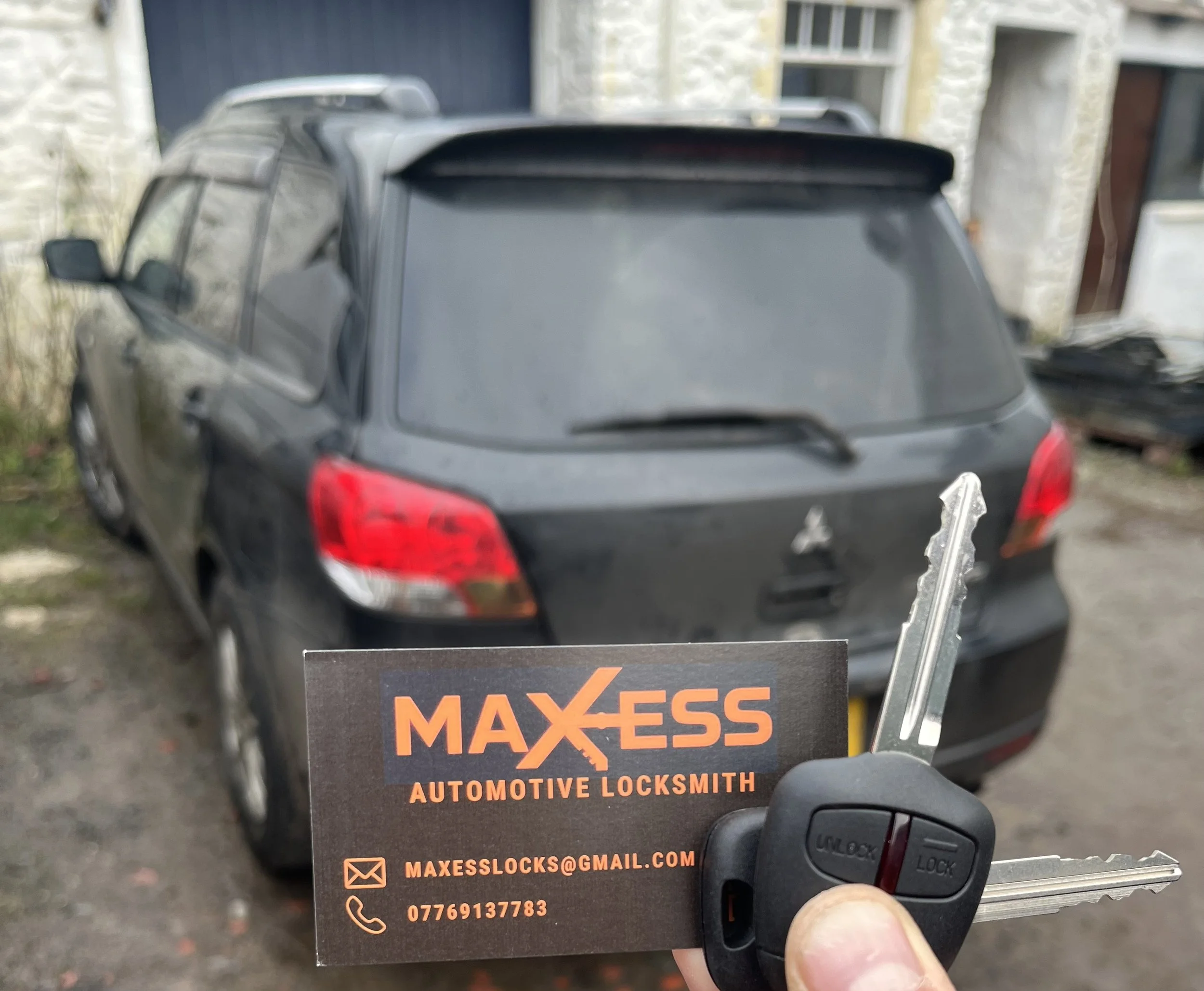 A black hatchback car parked outside a building with a dust-covered rear window. The person in the image is holding a car key and business card for Maxess Automotive Locksmith, with contact information visible.