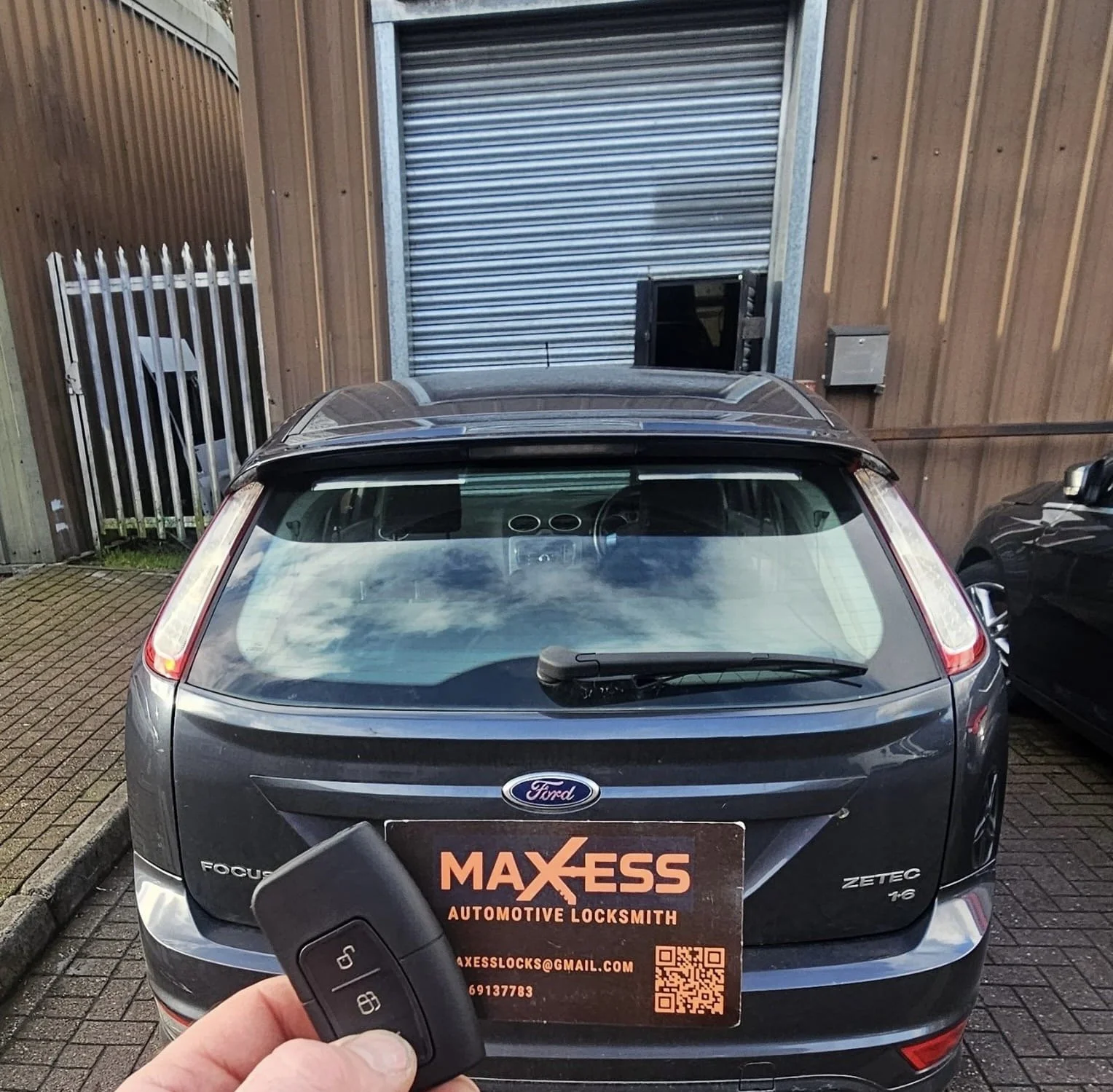 The image shows the rear view of a black Ford Focus Zetec parked outdoors near a wooden building with a metallic roll-up door. A person in the foreground holds a car key and an automotive locksmith business card that reads 'MAXESS AUTOMOTIVE LOCKSMIT