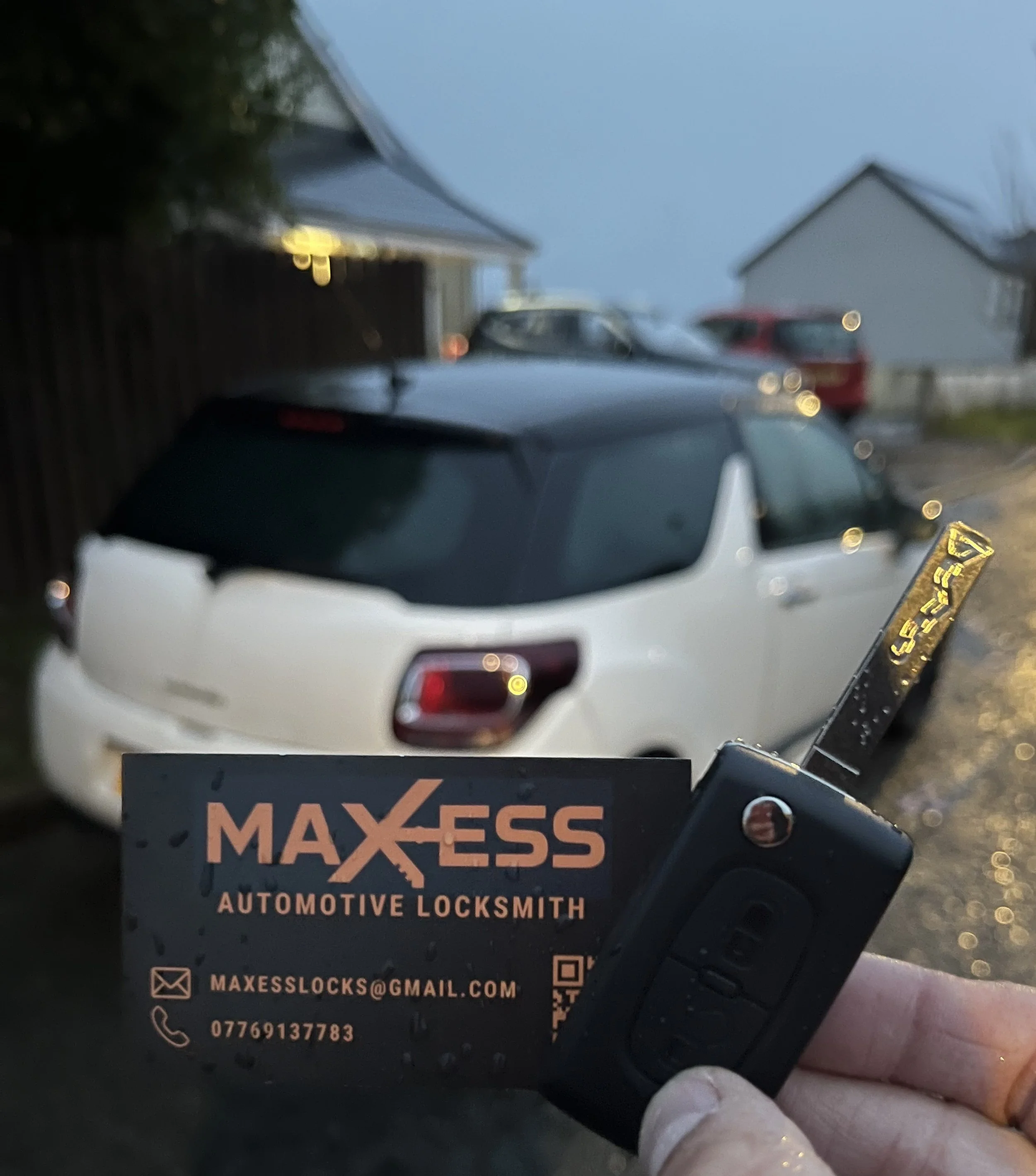 Person holding an automotive locksmith business card and car key in front of a white car parked outside during dusk.