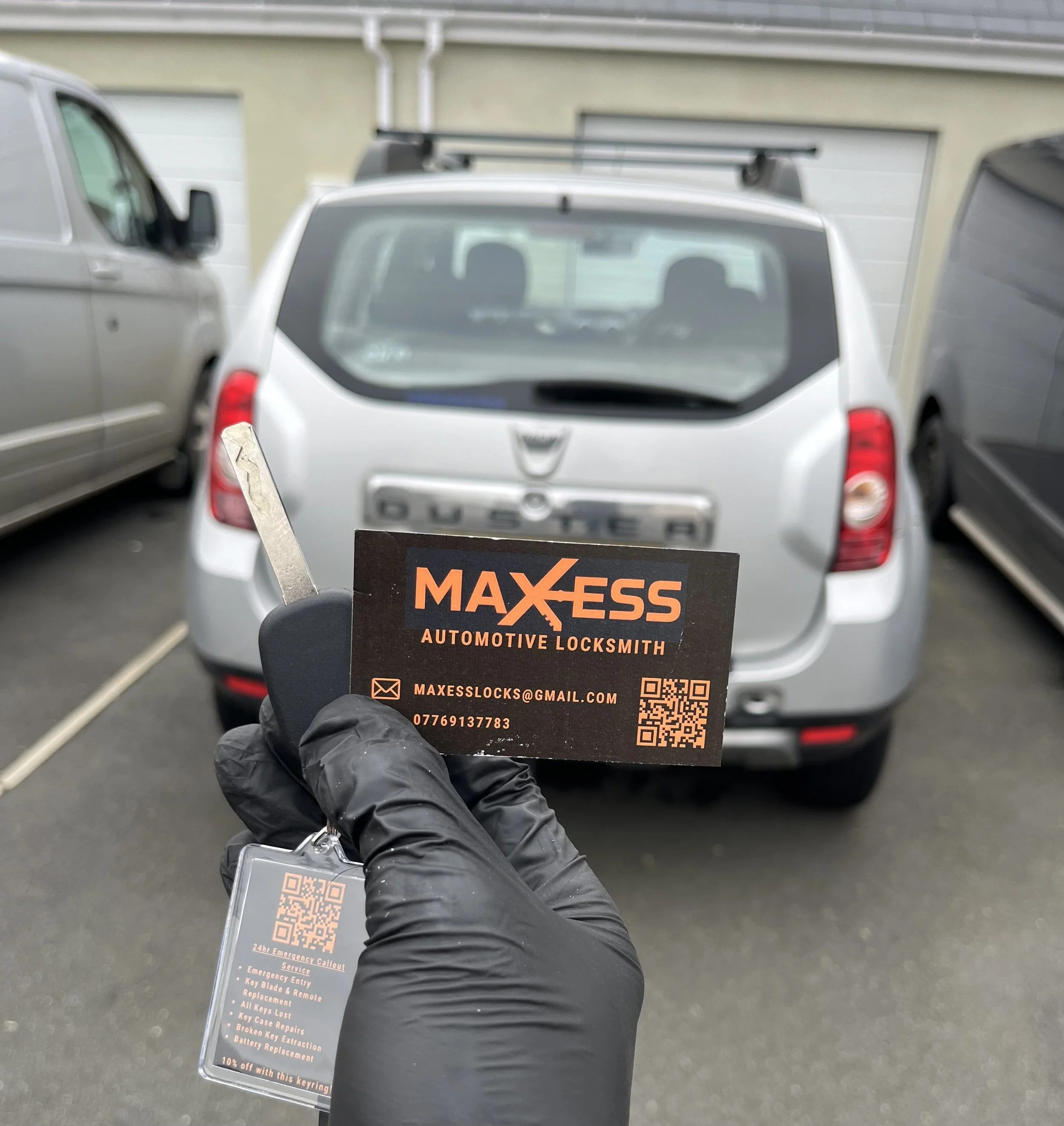 Hand wearing a black glove holding car keys and a business card for Maxess Automotive Locksmith in front of a silver Dacia Duster parked in a driveway.