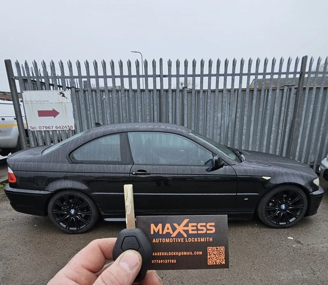A black BMW coupe parked in front of a metal fence, with a hand holding a car key and a business card for Maxess Automotive Locksmith in the foreground.