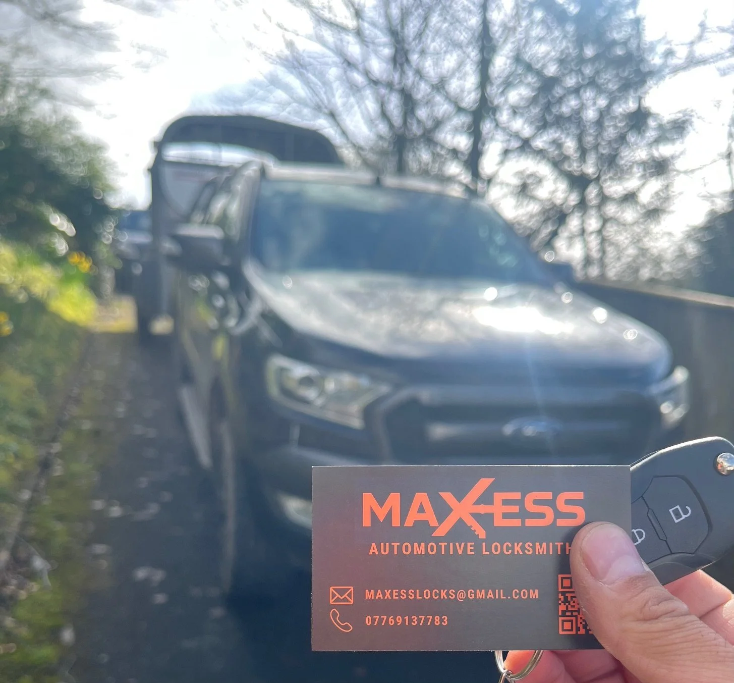 sHand holding a business card for MAXESS Automotive Locksmith in front of a black SUV on a farm road. Showing a replacement car key