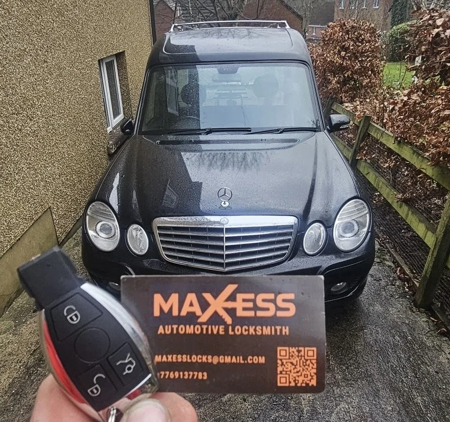 A black Mercedes-Benz car parked in a narrow area between a building and a wooden fence, with a person's hand holding a key fob and business card for Maxess Automotive Locksmith in front of it.