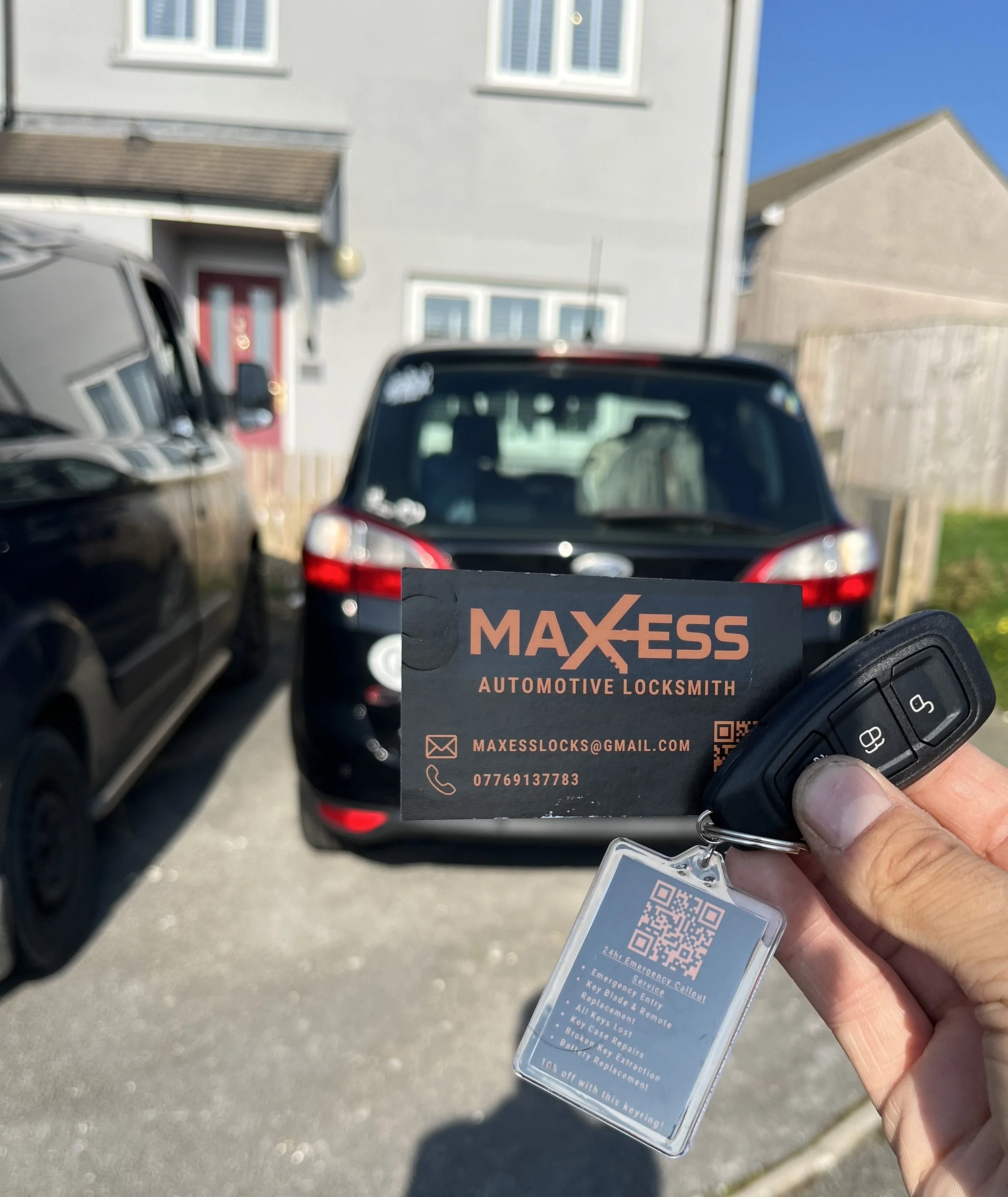 A person holding a car key next to a business card for Maxess Automotive Locksmith with a QR code. Car parked in the background, with other vehicles and residential buildings visible.