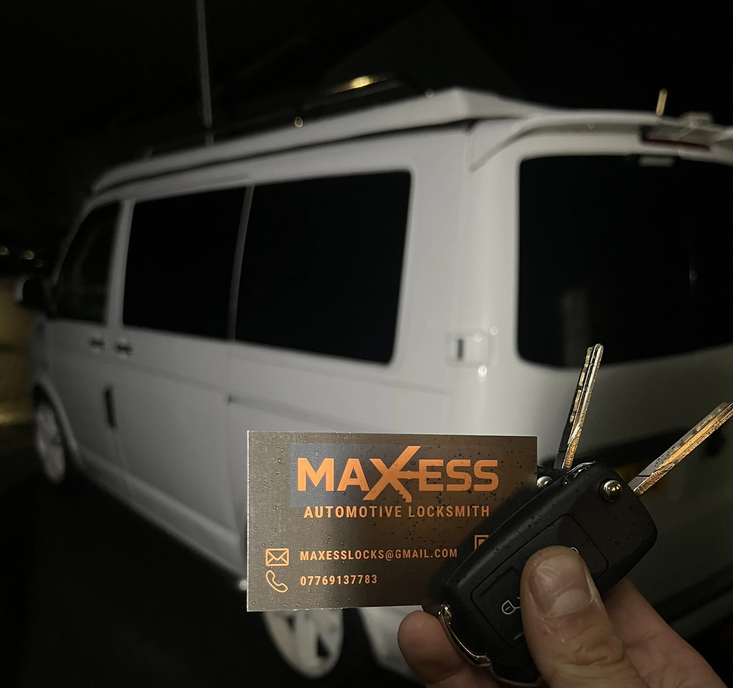 A person holding a car key and a business card for Maxess Automotive Locksmith in front of a white van at night.
