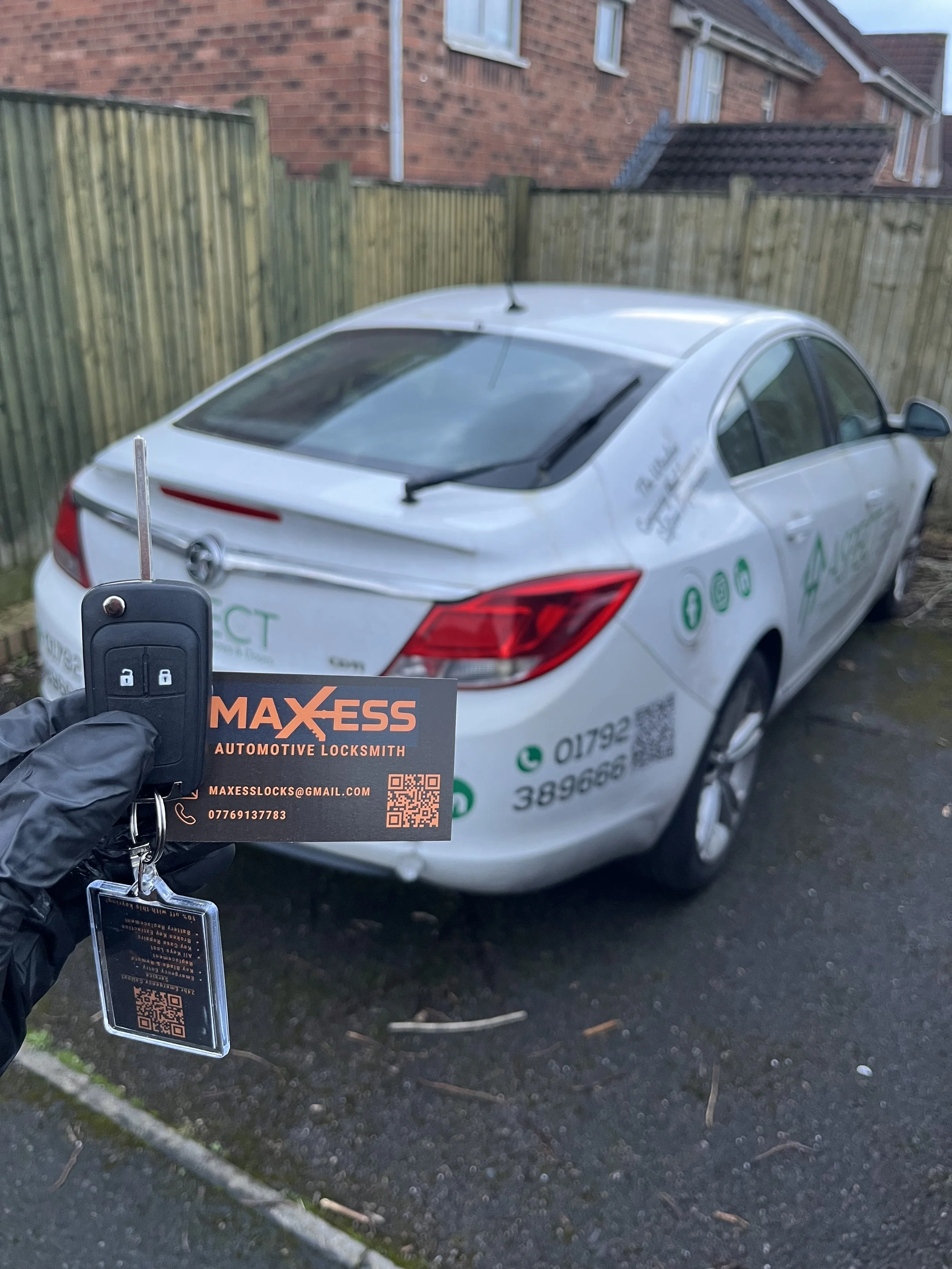 A white car parked on a driveway next to a wooden fence. A person wearing a black glove holds a car key and a business card for MAXESS Automotive Locksmith in front of the car. The car has decals and contact information on its side.