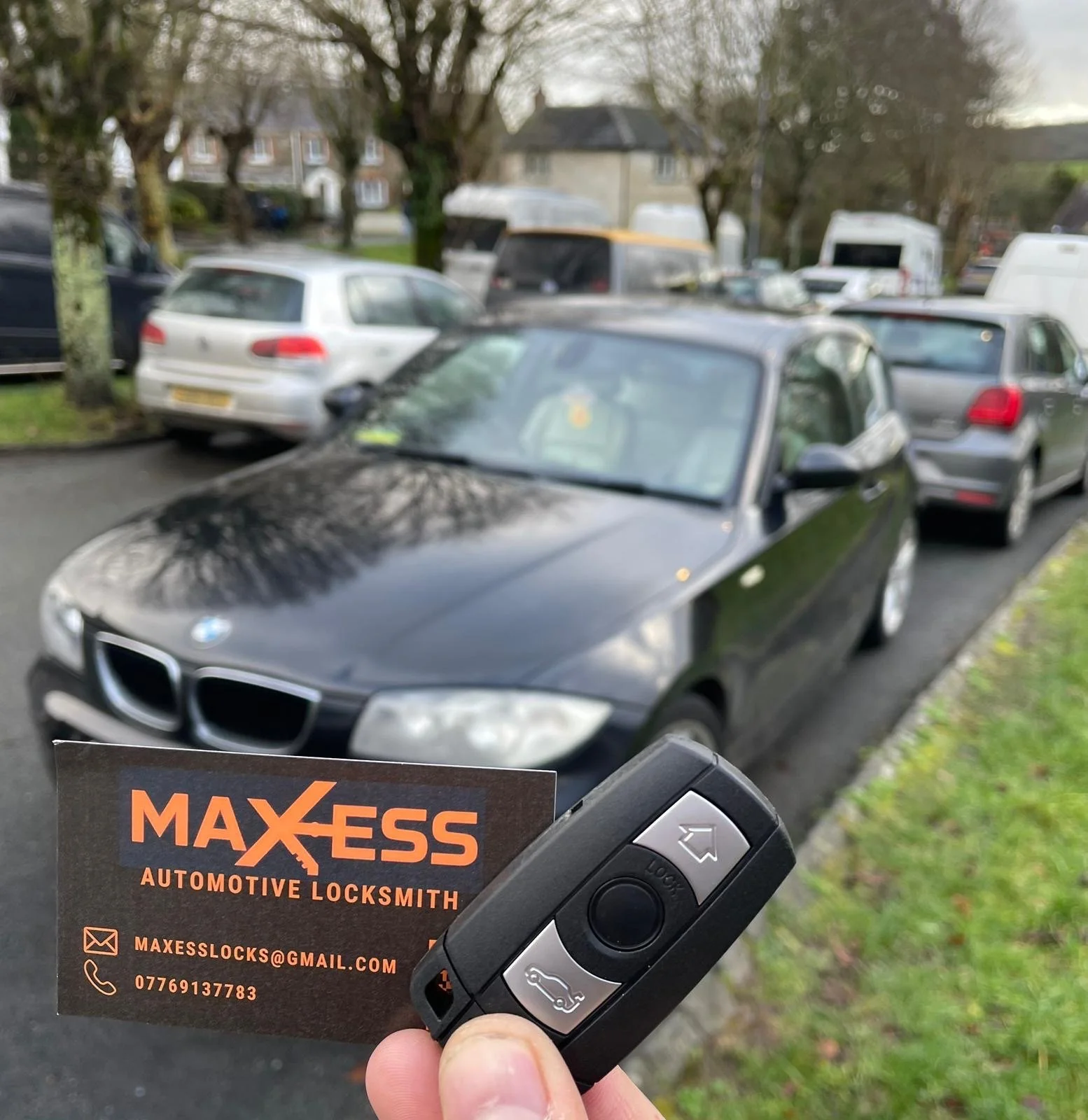 A person holding a car key and a business card in front of a black BMW parked on a street with other vehicles and trees in the background.
