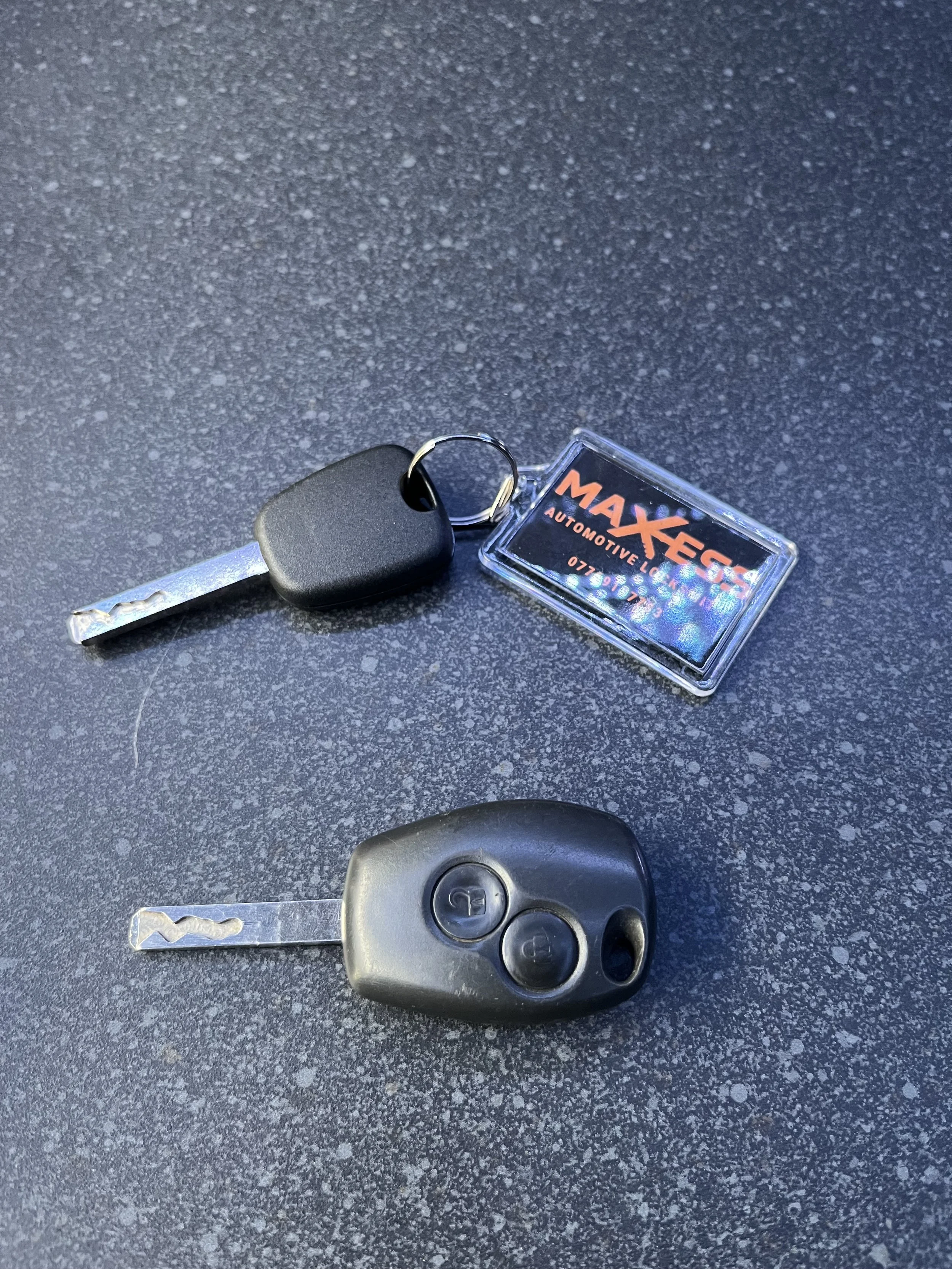 A set of car keys on a black textured surface, including a remote key fob and a key with a black plastic head, and a small plastic keychain with the orange and red MAXECS logo.