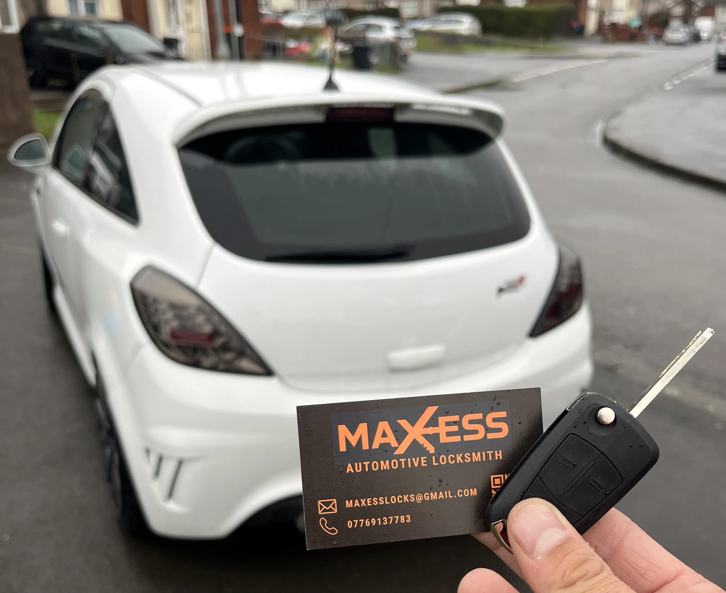 Person holding a car key and a business card for Maxess Automotive Locksmith with a white hatchback car in the background on a street