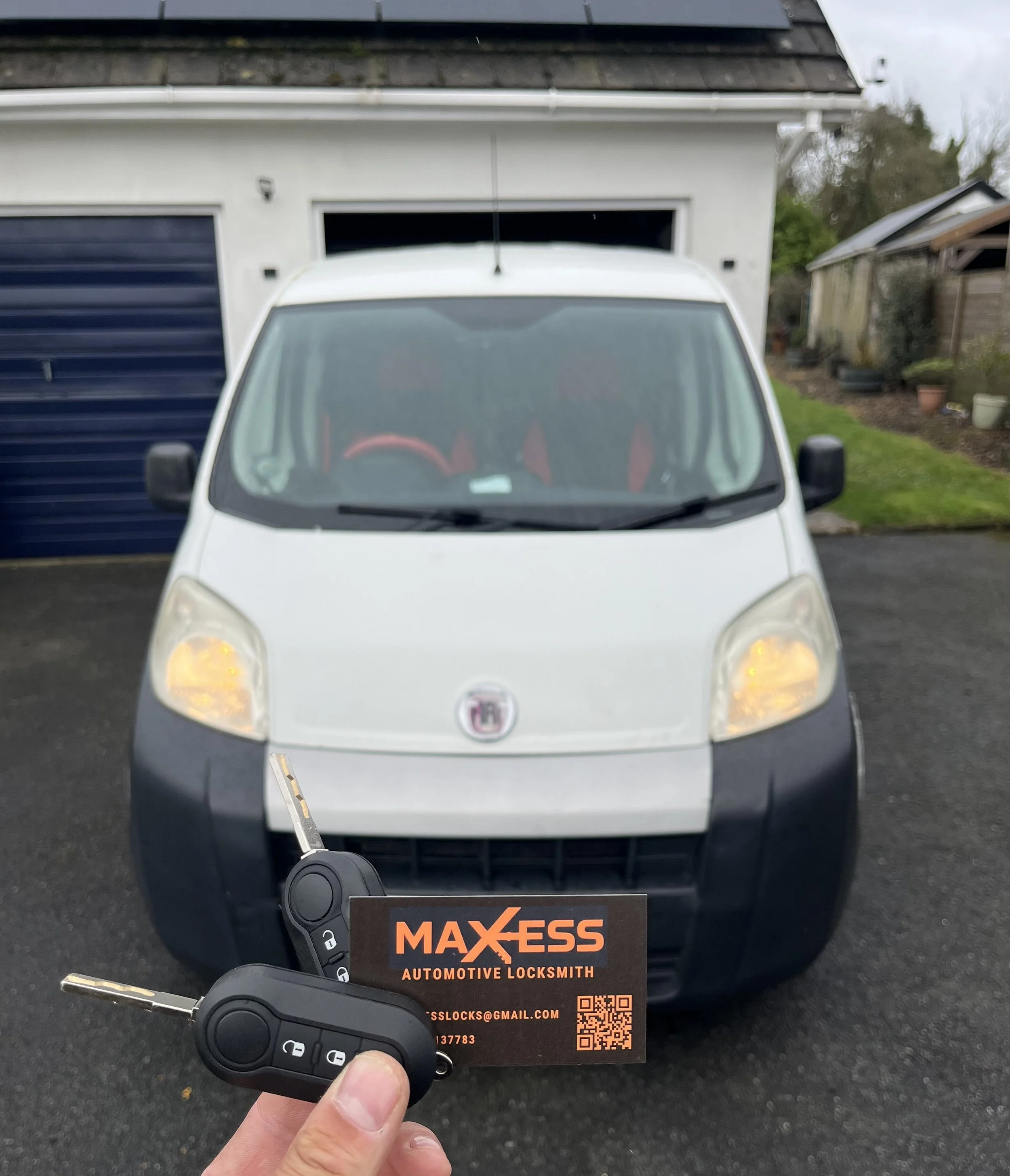 A white van parked in front of a garage with a black door. A hand holding two car keys and a business card for MAX-ESS Automotive Locksmith in the foreground.