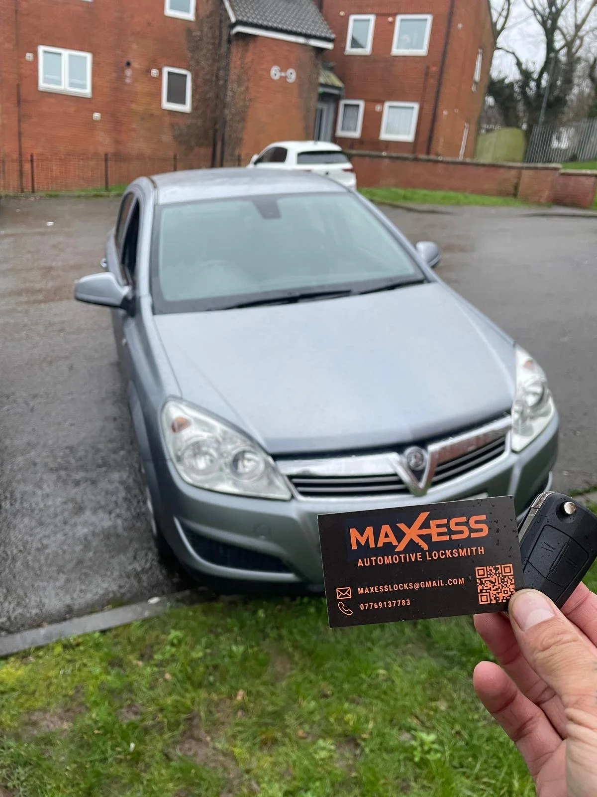 A person holding a car key and business card in front of a silver car in an outdoor parking lot, with a brick residential building in the background.