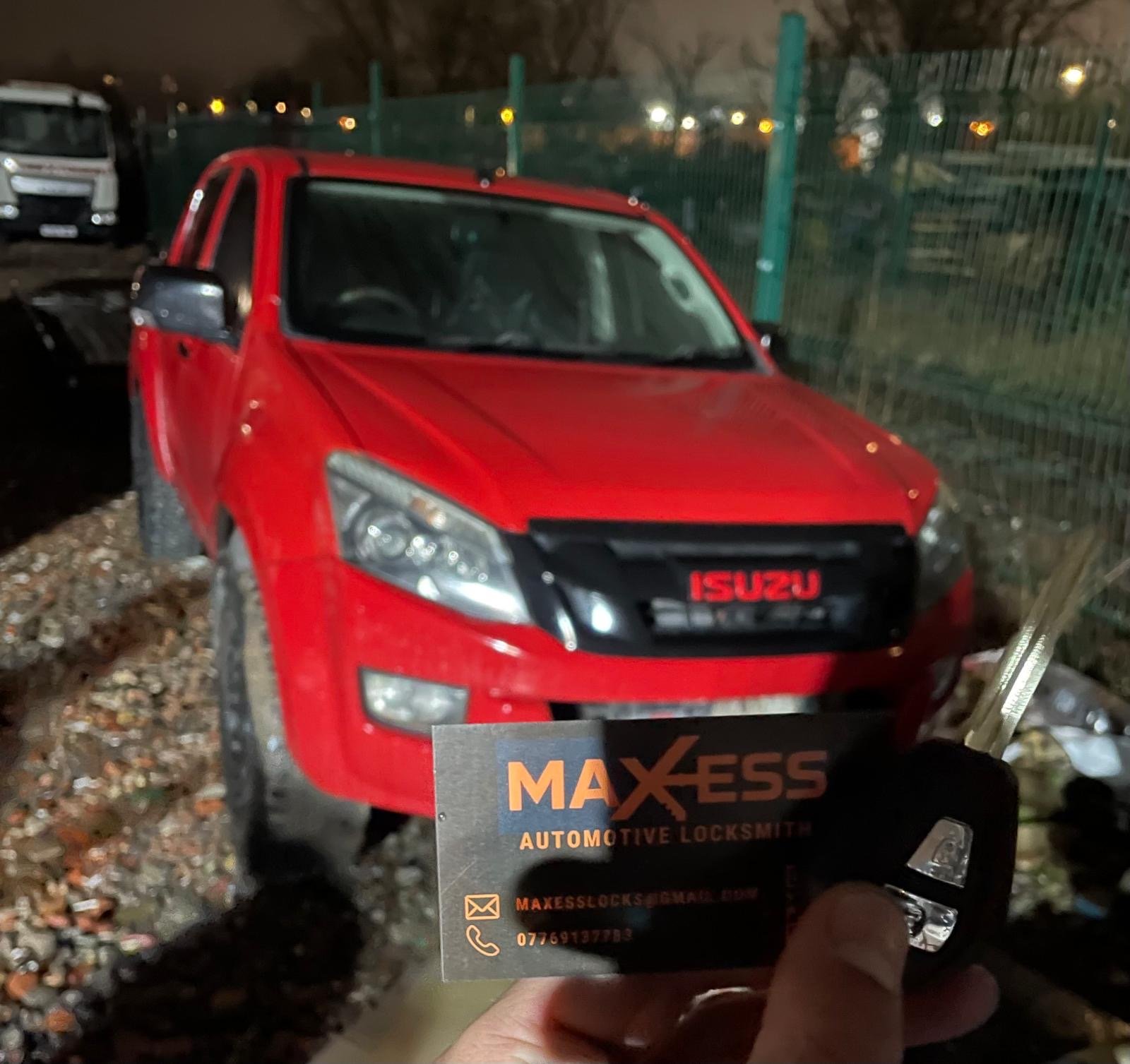 A red Isuzu pickup truck parked outdoors at night, with a business card for Maxess Automotive Locksmith held in the foreground.