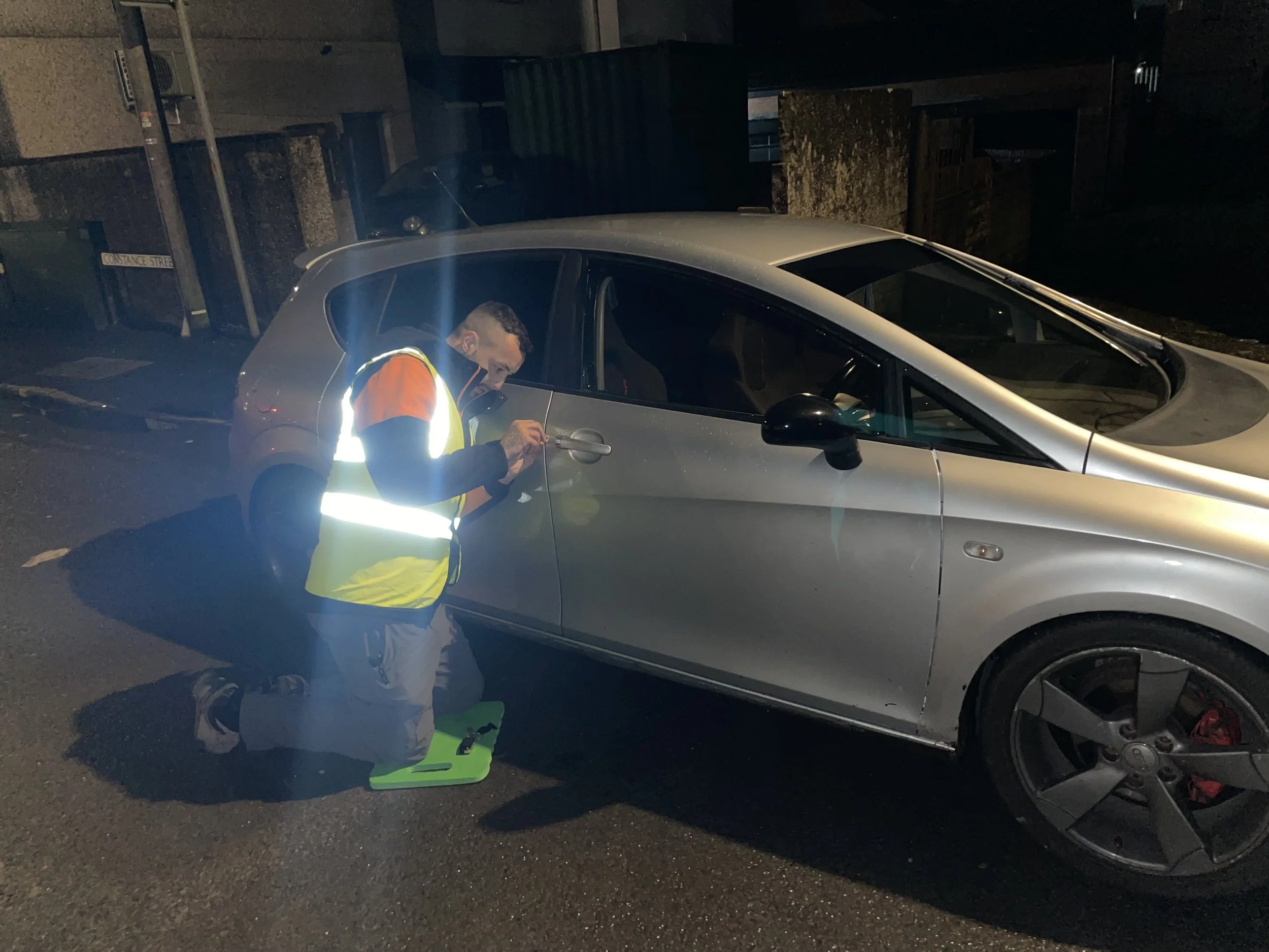 A man wearing a high-visibility safety vest kneeling on the road next to a silver car at night, inspecting or working on the car's door handle with a tool in his hand.