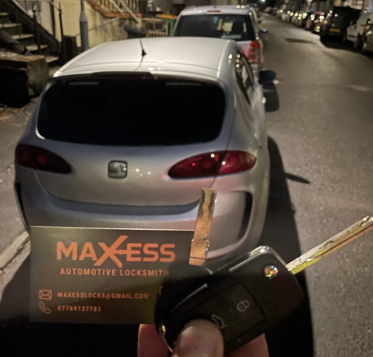 A person holding a key fob and a business card for MAXXESS Automotive Locksmith, with a silver car parked on a city street at night in the background.