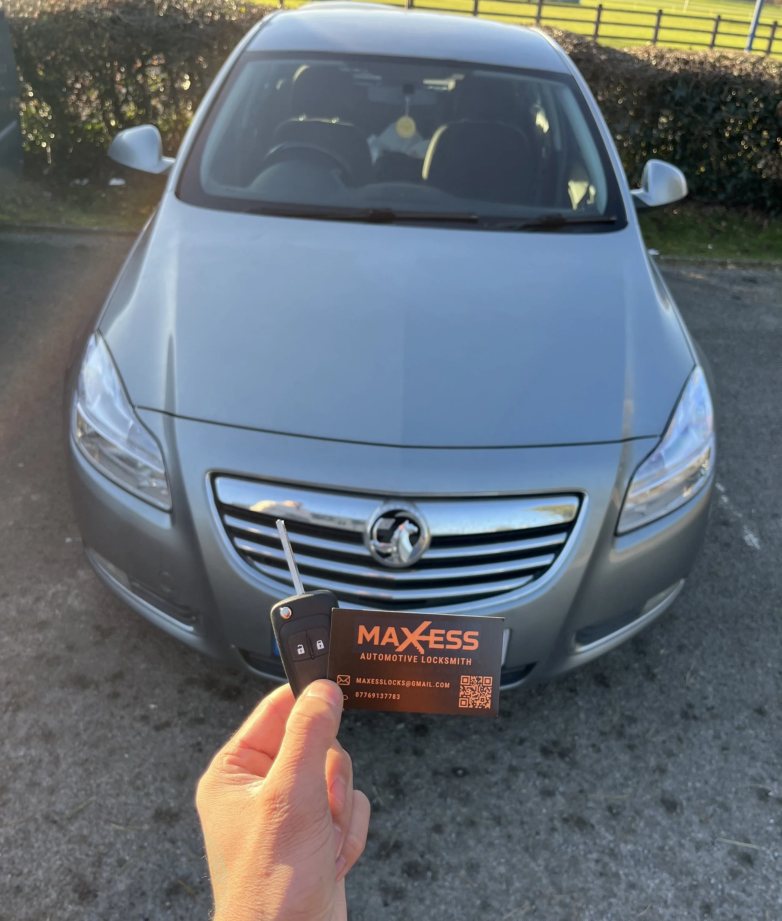 Silver Holden sedan parked on asphalt, held in front of the camera by a hand holding a car key and business card from Maxess Automotive Locksmith.