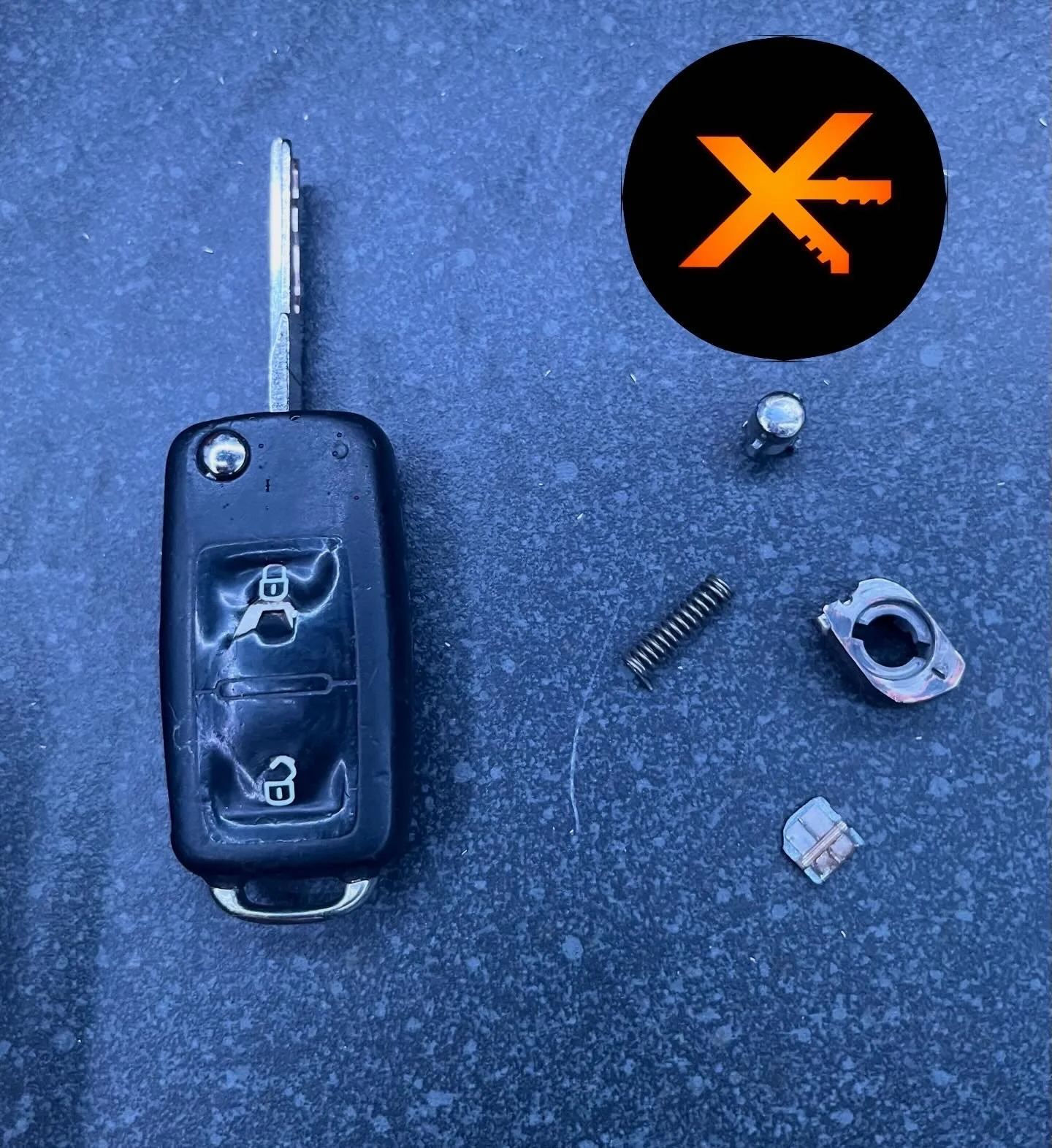 Disassembled car key with broken remote buttons and various small metal parts on a dark textured surface, with a black and orange warning icon in the upper right corner.