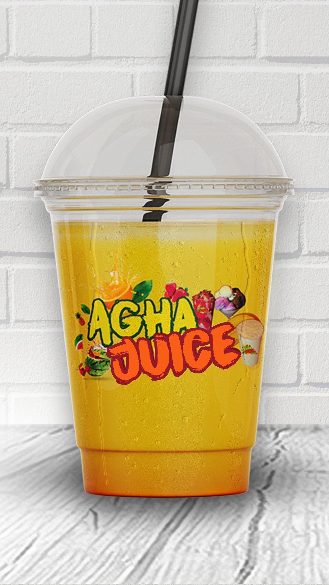 A clear plastic cup with yellow-orange liquid and a black straw, labeled 'Aloha Juice' with tropical fruit graphics, set against a white brick wall and wooden surface.