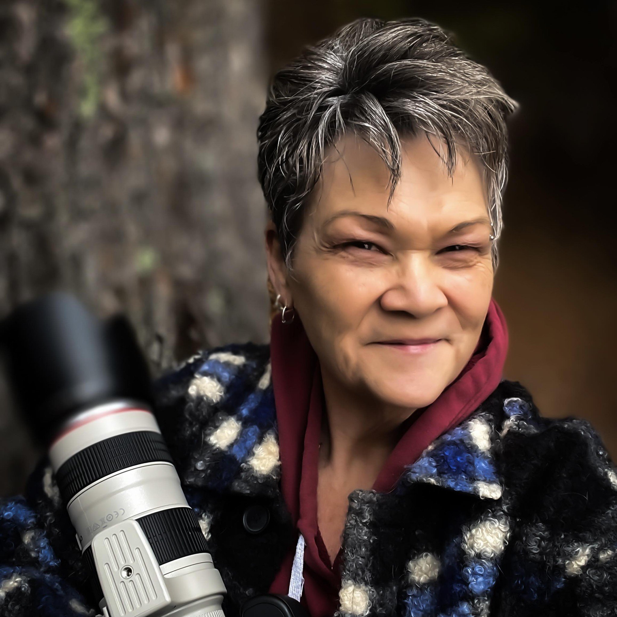 Smiling woman with short, gray hair holding a camera lens, wearing a multicolored patterned coat and a maroon hoodie, standing outdoors near a stone wall.