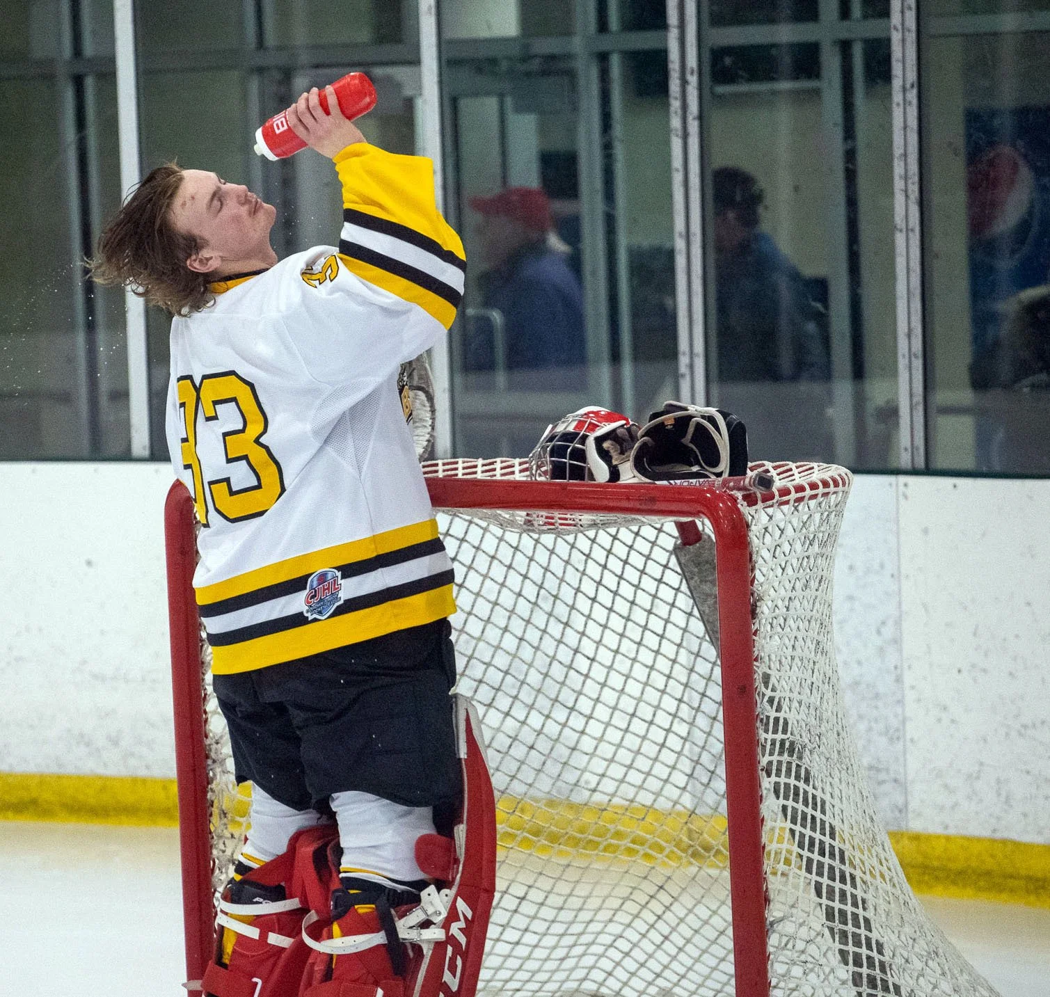 Young Smiths Falls Jr. A Bears' hockey goalie in a white and black jersey with yellow accents, standing by the goal post, holding a water bottle, with helmet and gloves placed on the net, in an ice hockey rink.