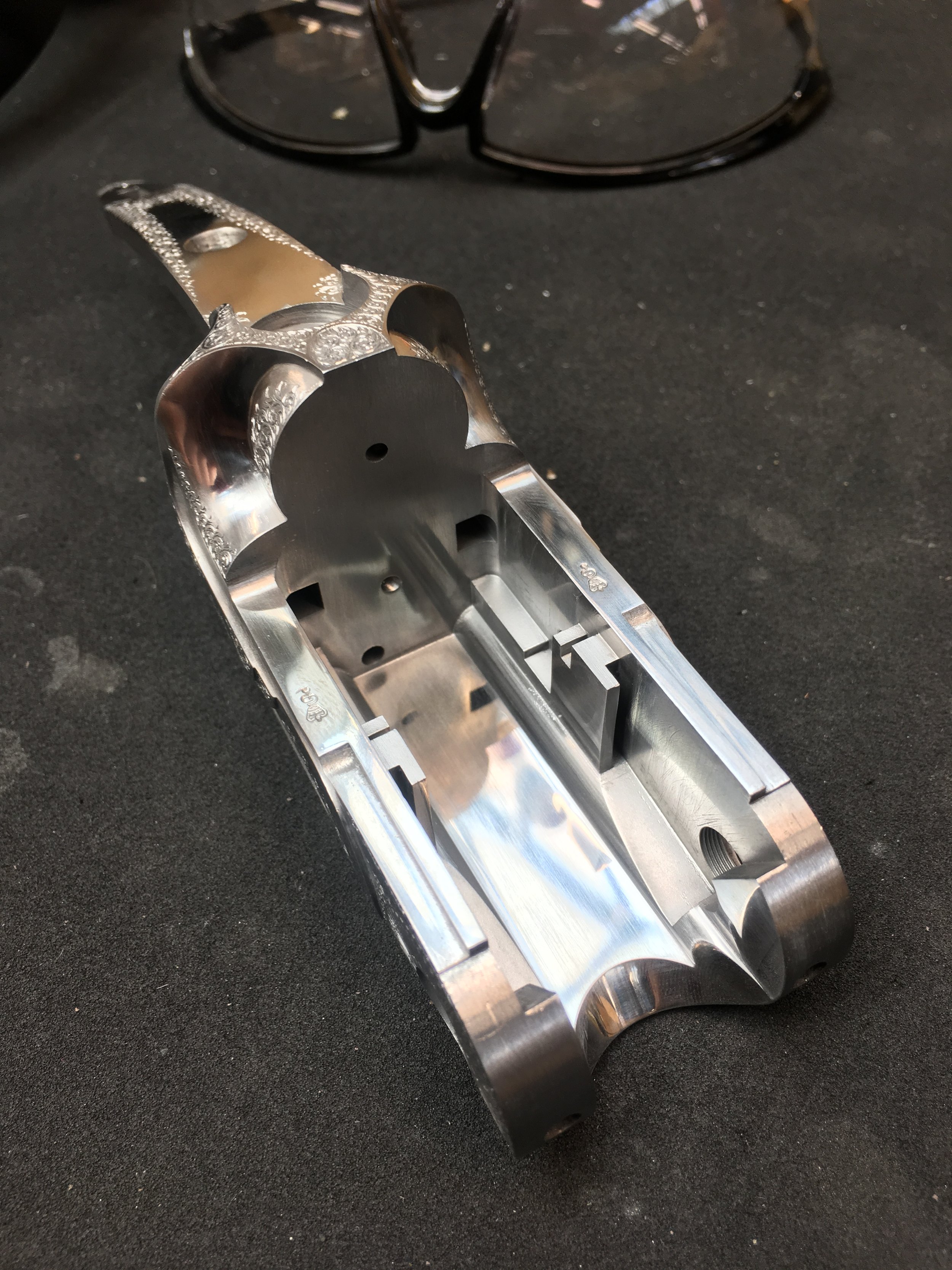 A shiny, metallic, and intricately machined gun frame or receiver with some glasses in the background on a black surface.