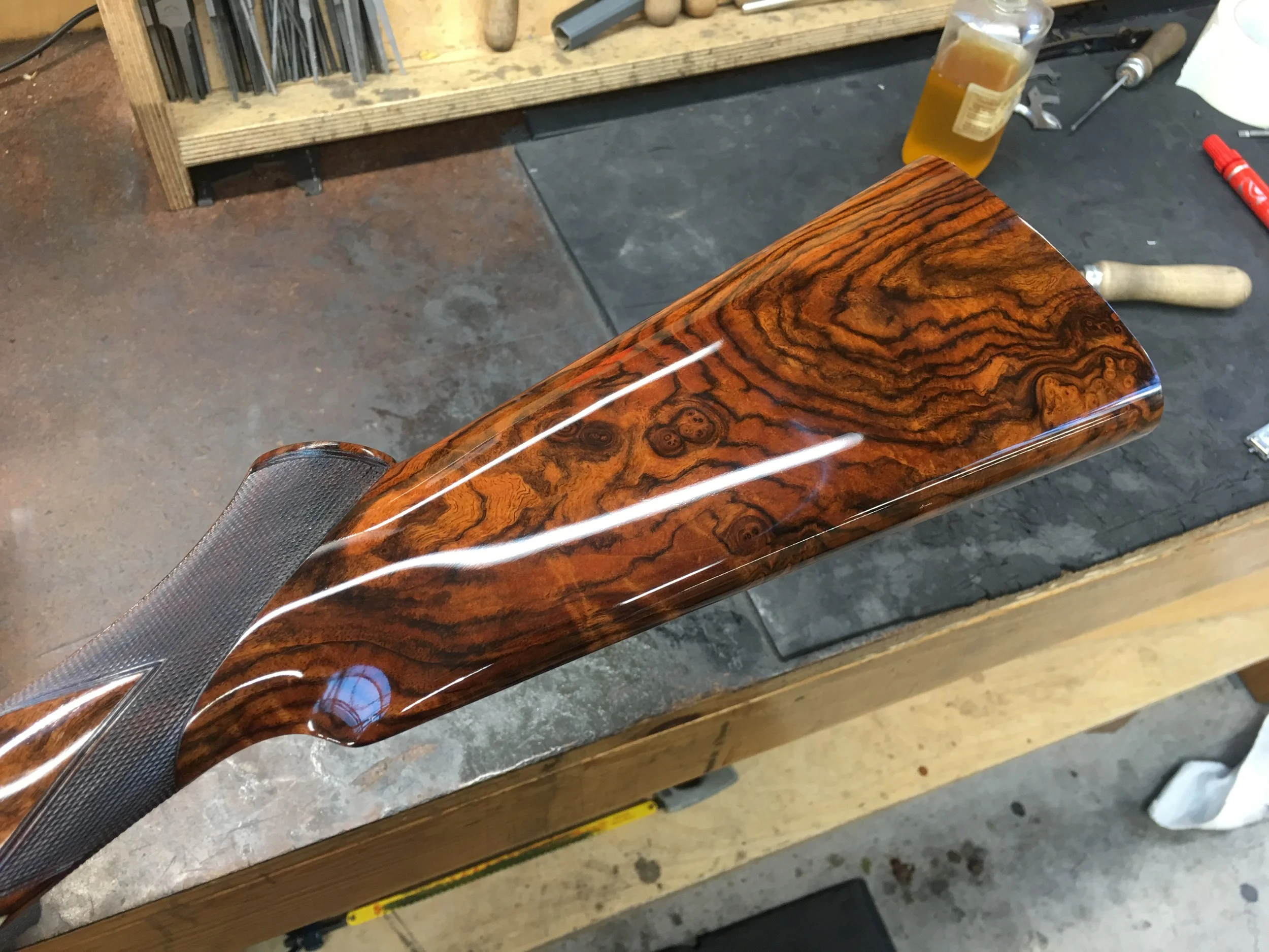 Close-up of a wooden rifle stock in a workshop, with tools and a bottle of oil in the background.
