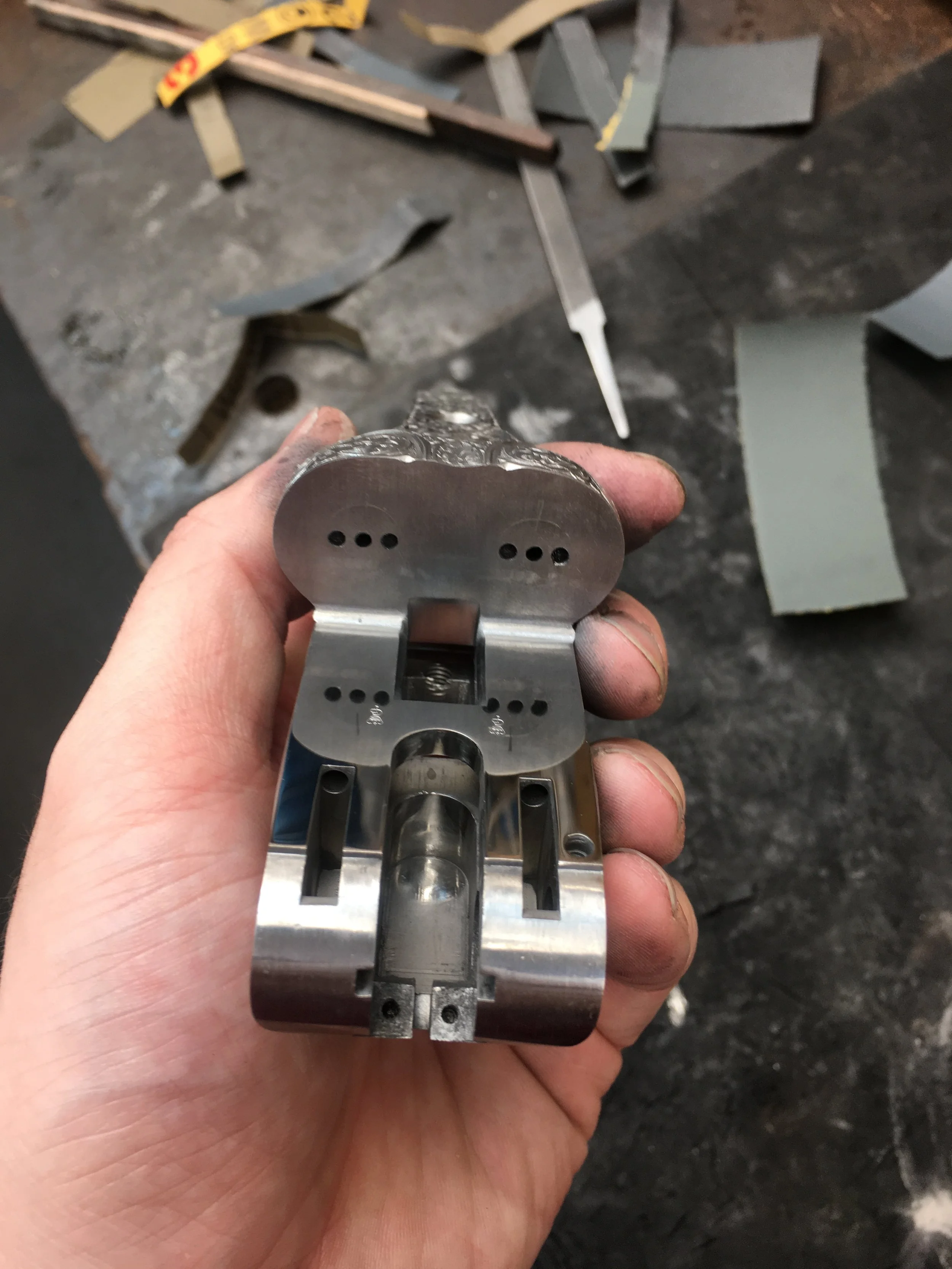 A close-up of a metal mechanical component being held by a person's hand, with a workshop or fabrication environment in the background, including tools and metal scraps.