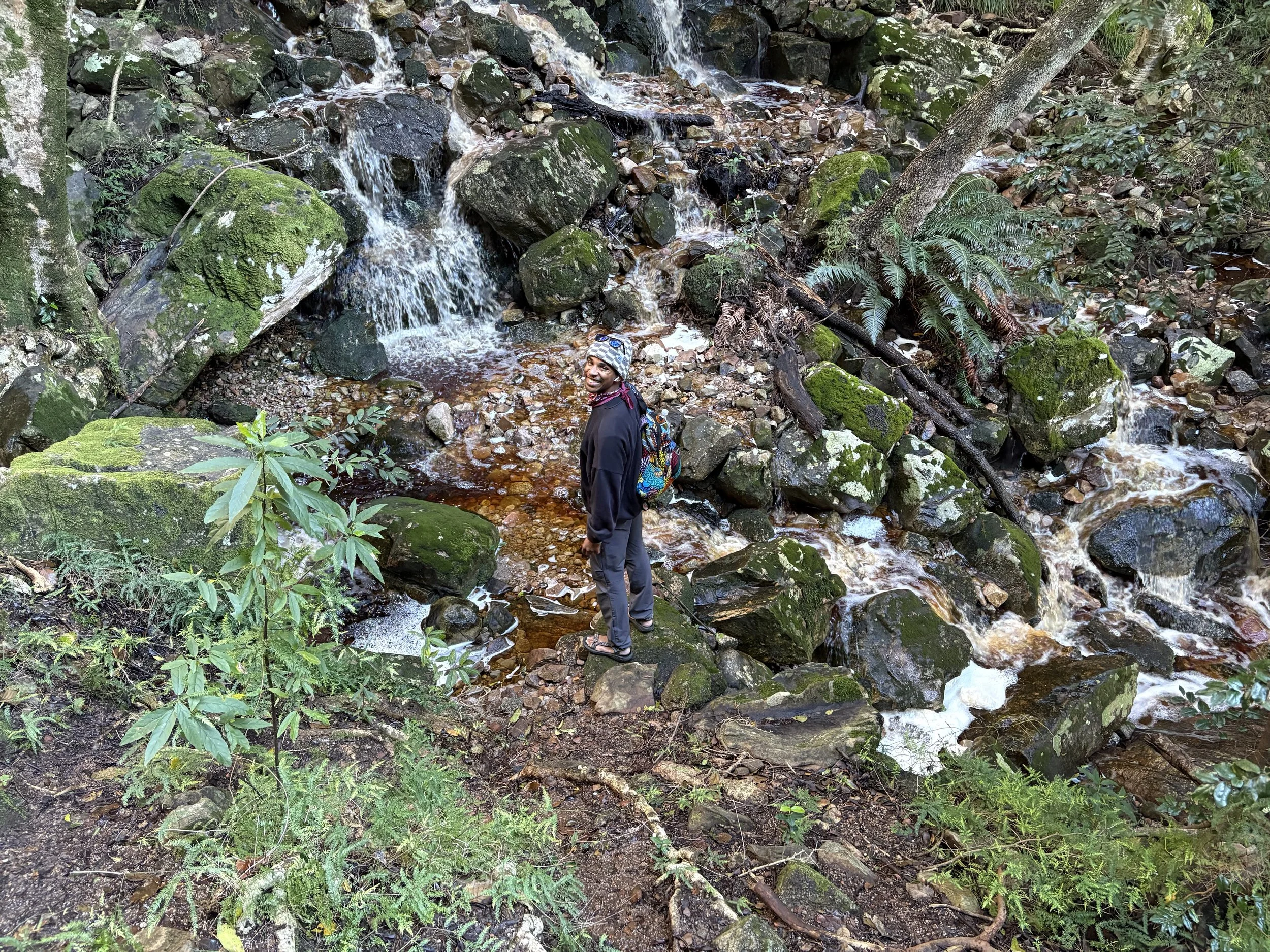Person standing on rocks near a small waterfall in a lush forest.