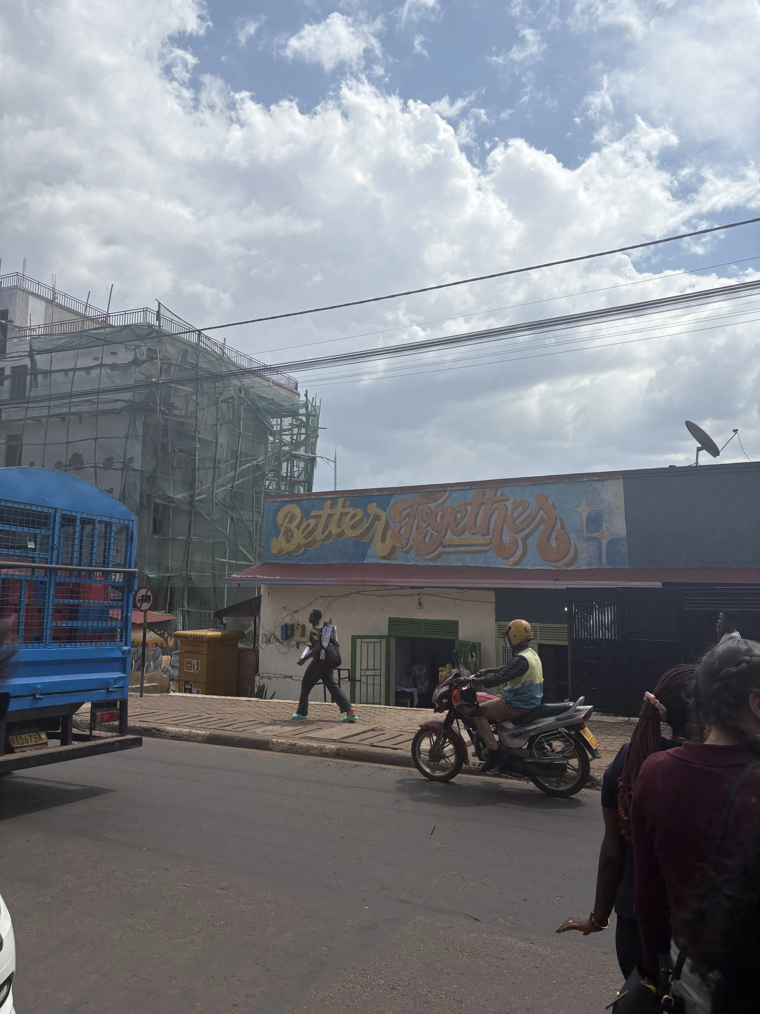 Street scene with pedestrians and a motorcyclist passing by a building with a mural that says 'Better Together,' a construction site, and a partly cloudy sky.