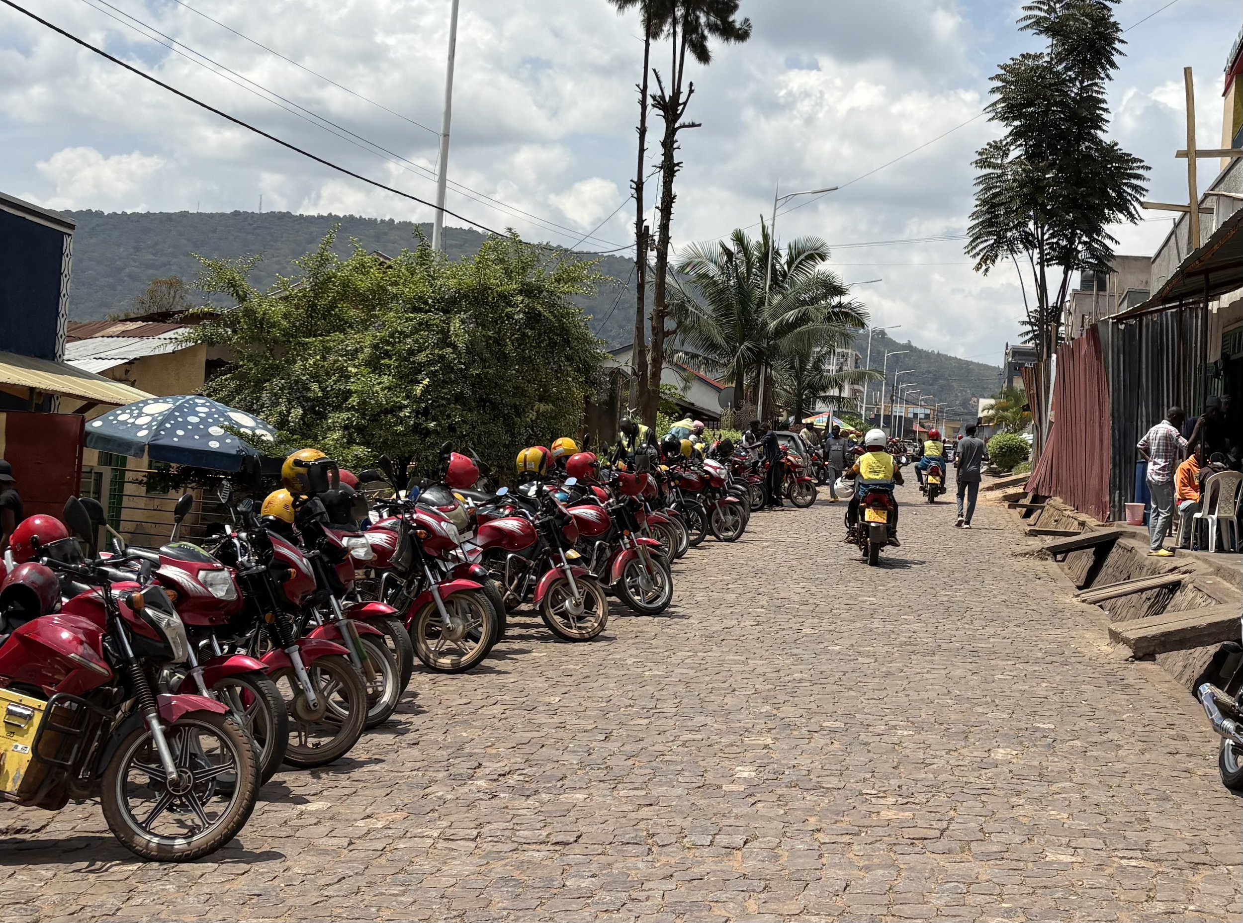 A cobblestone street in a small town with numerous red motorcycles and helmets parked on the left side. People are walking and riding motorcycles along the street, which is lined with trees and simple buildings, with hills in the background and a par