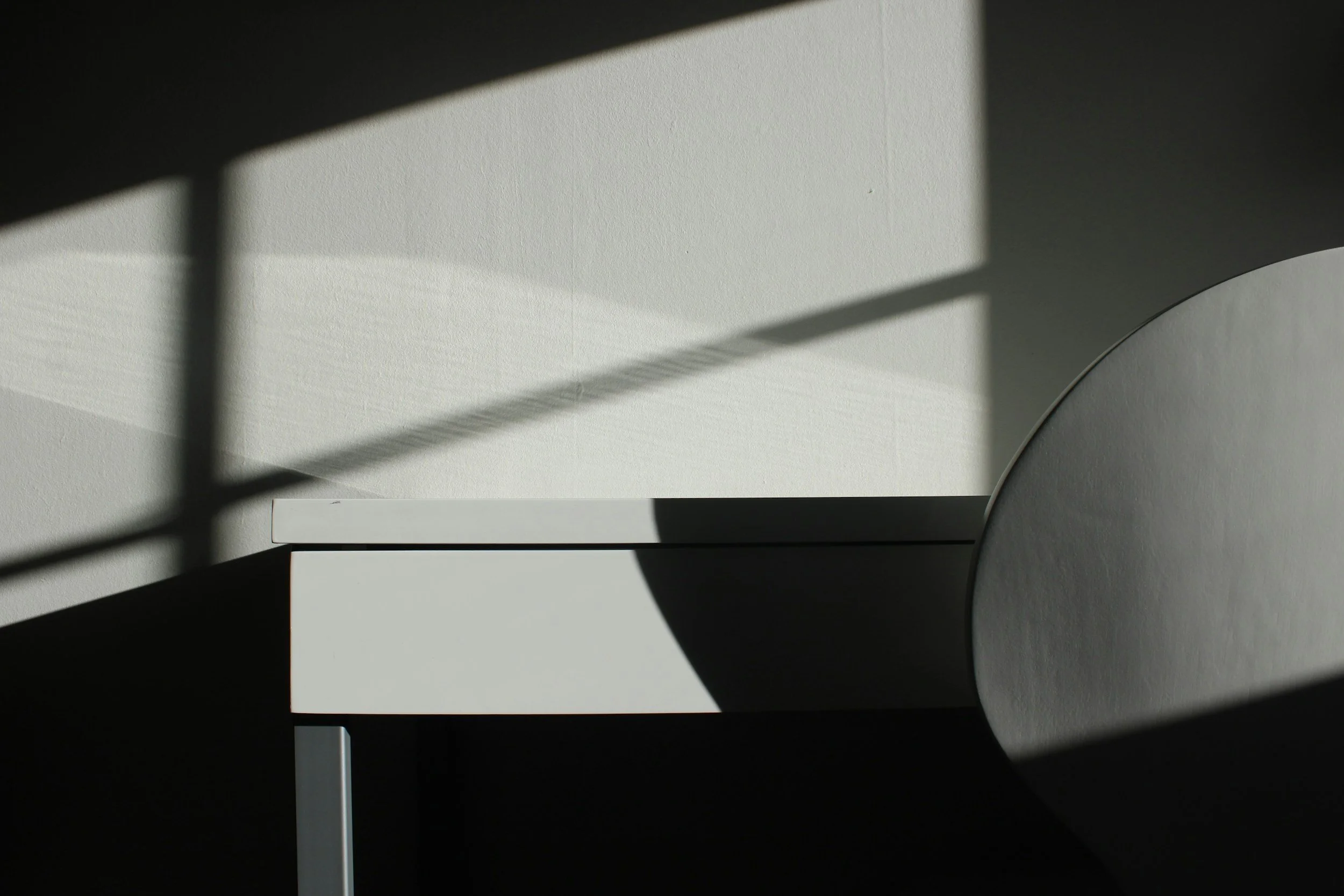A minimalist corner of a room with a gray table or desk, white wall with textured surface, and geometric light and shadow patterns cast on the wall and table.