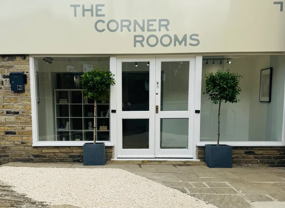 The entrance to a business named 'The Corner Rooms' with a glass door, two potted trees outside, and interior signage.
