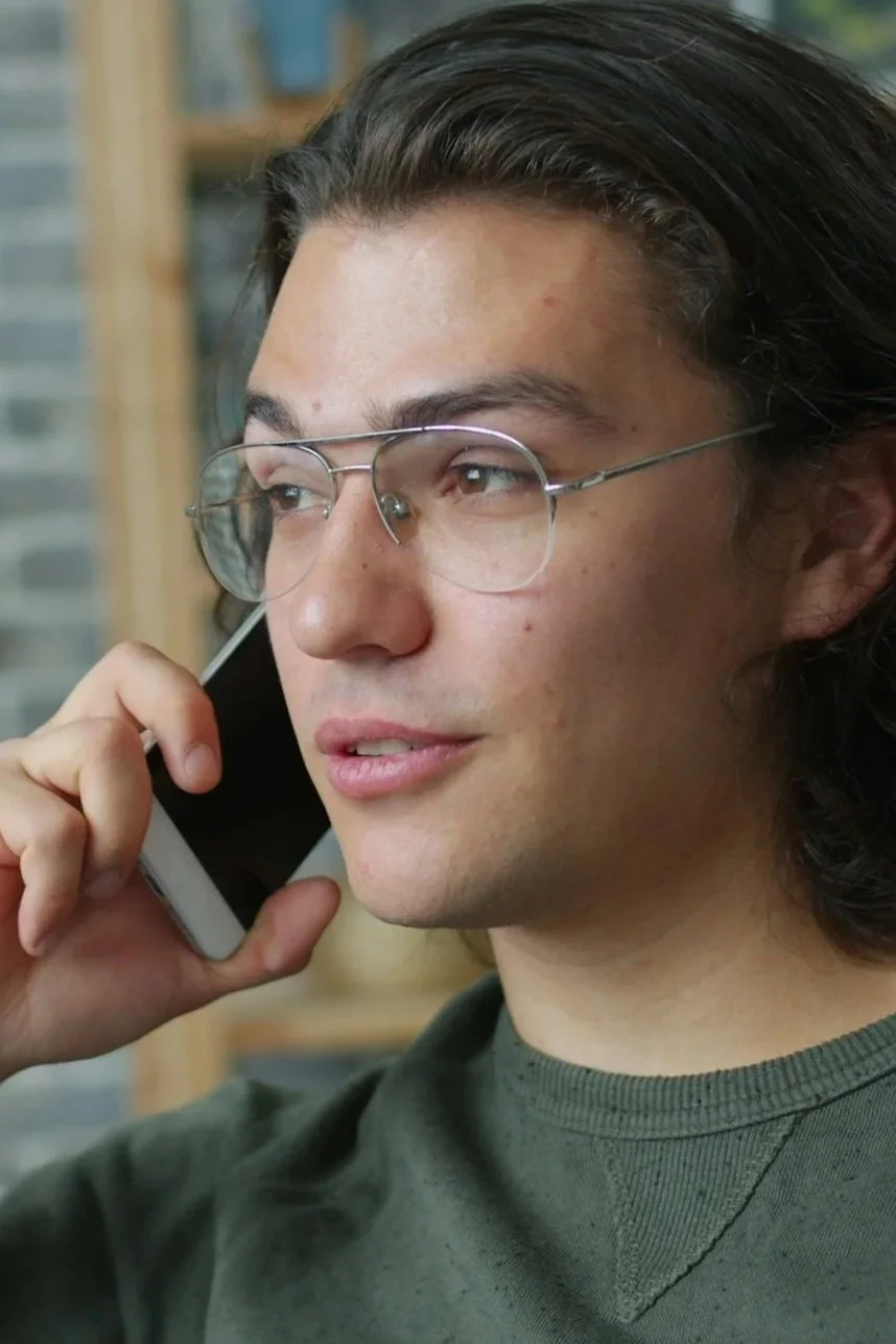 A young man with long dark hair and glasses is talking on a smartphone, with a blurred background of bookshelves.
