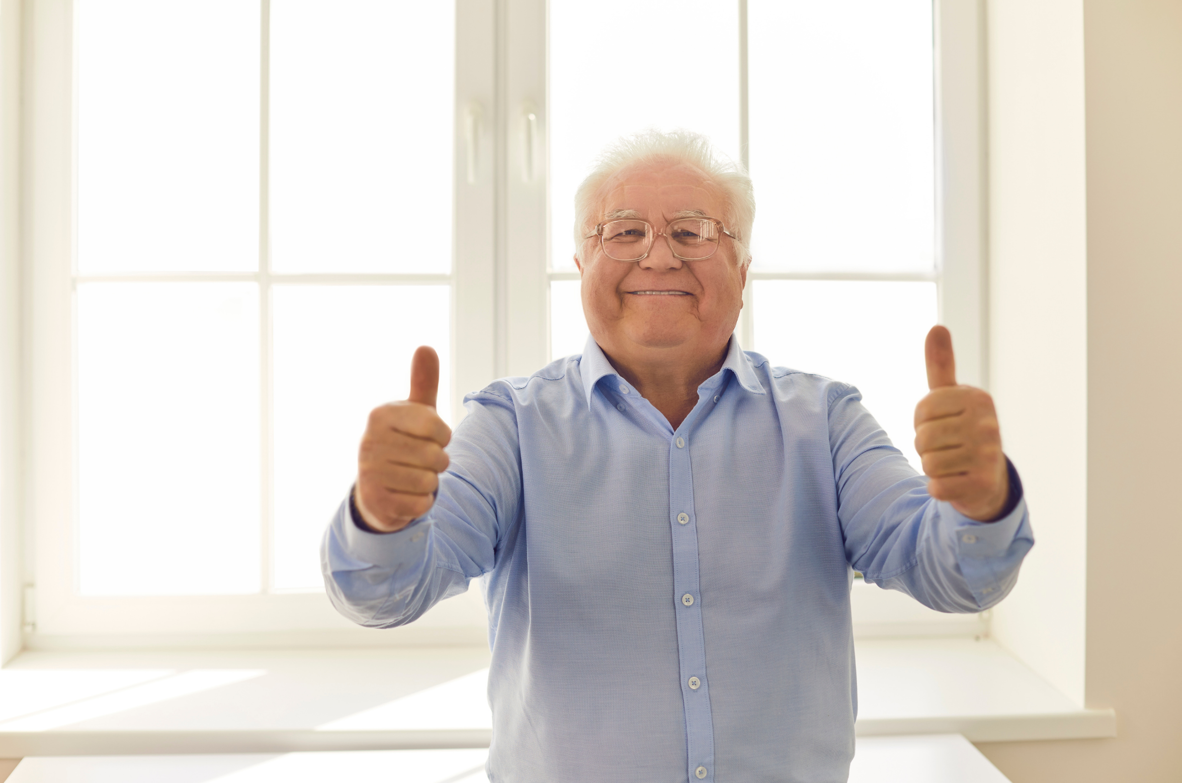 Un homme âgé, souriant, portant une chemise bleue, faisant un geste de pouce levé à chaque main dans une pièce lumineuse avec une grande fenêtre en arrière-plan.