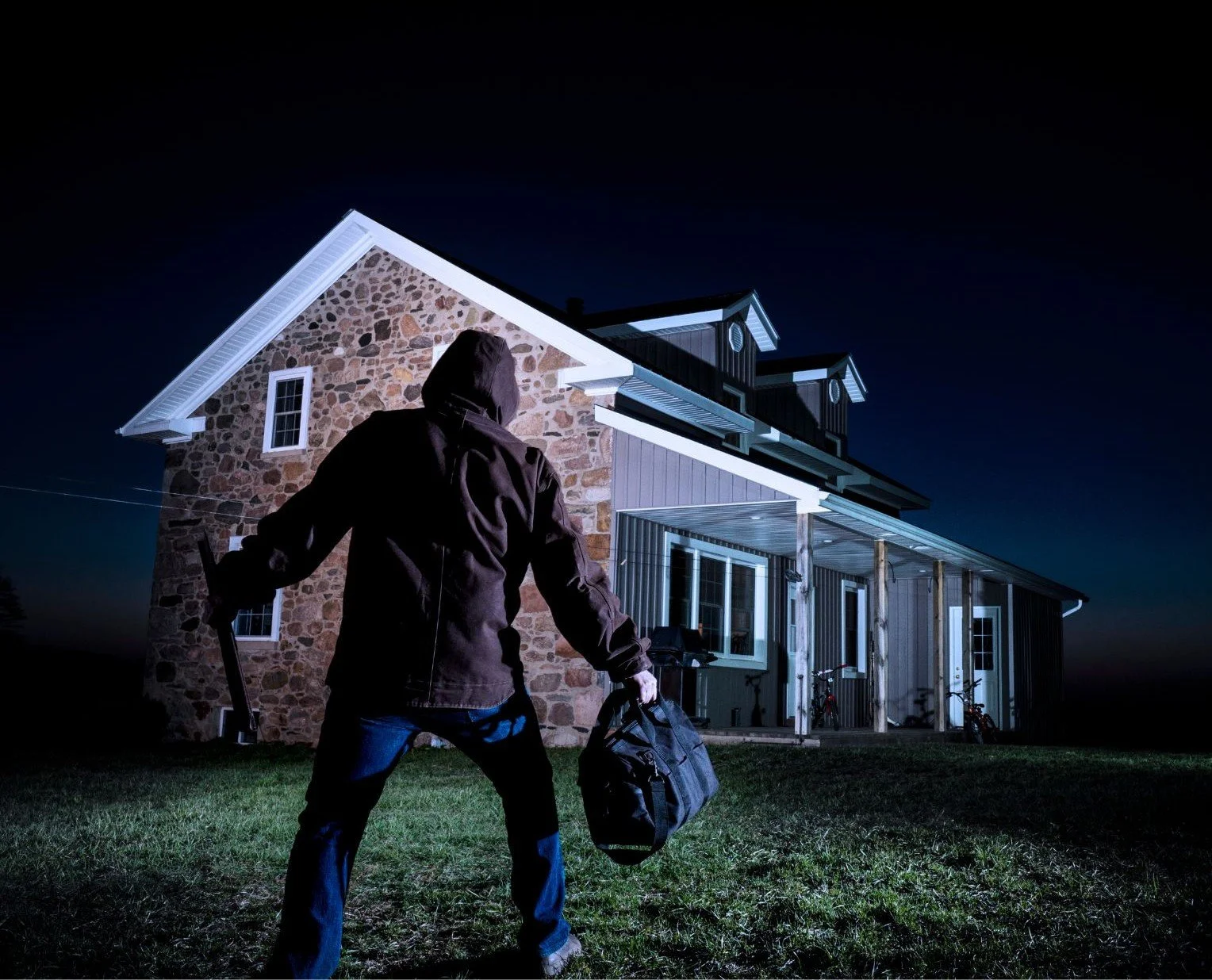 Une personne en manteau noir, de dos, tenant un sac pour cambrioler, se tient devant une maison en pierre essayant d'entrée