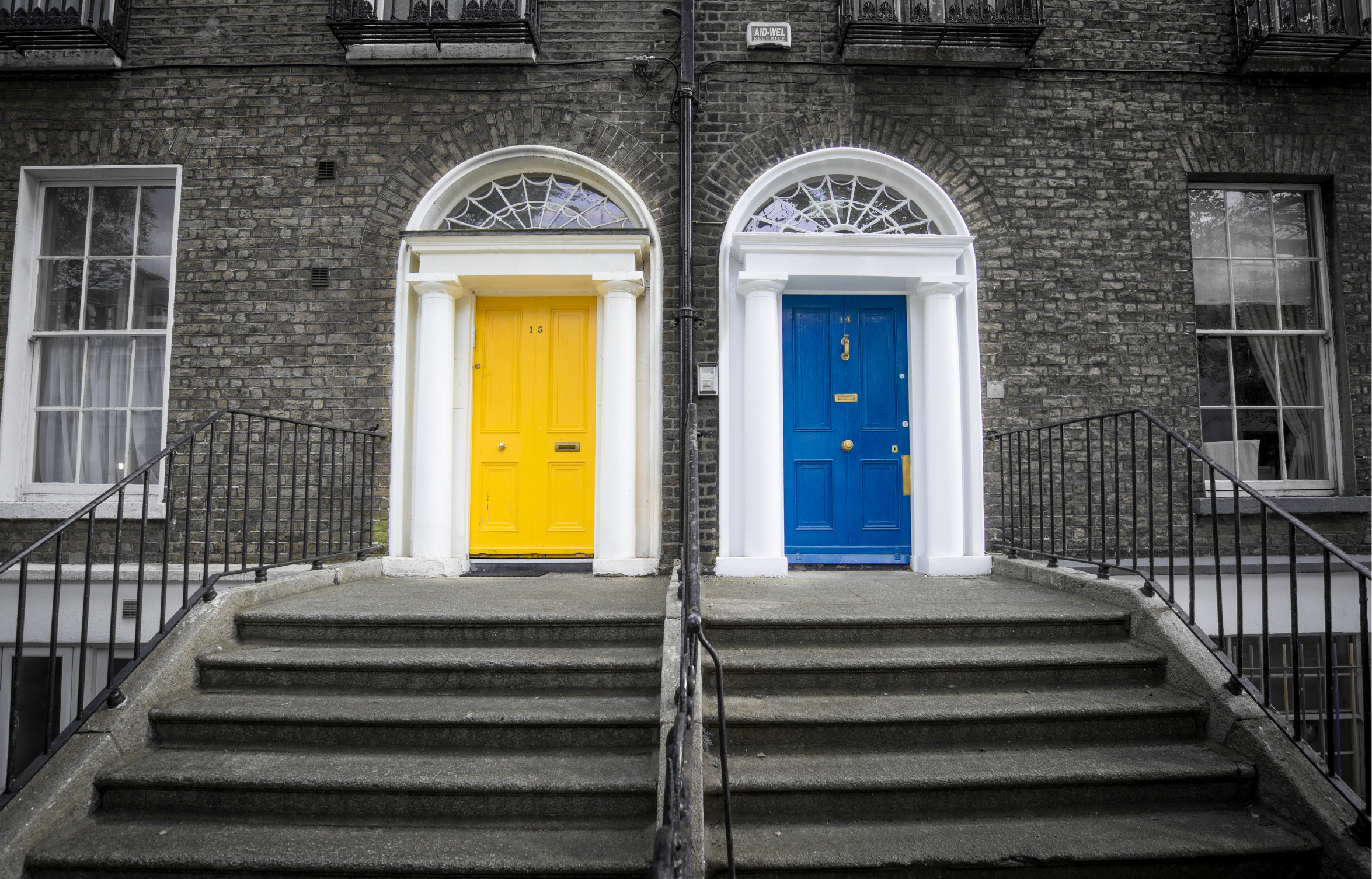 Deux portes d'entrée colorées, une jaune et une bleue, dans un bâtiment en brique avec des escaliers en béton et des rampes en métal, chaque porte encadrée de colonnes blanches et surmontée d'une fenêtre arqué.