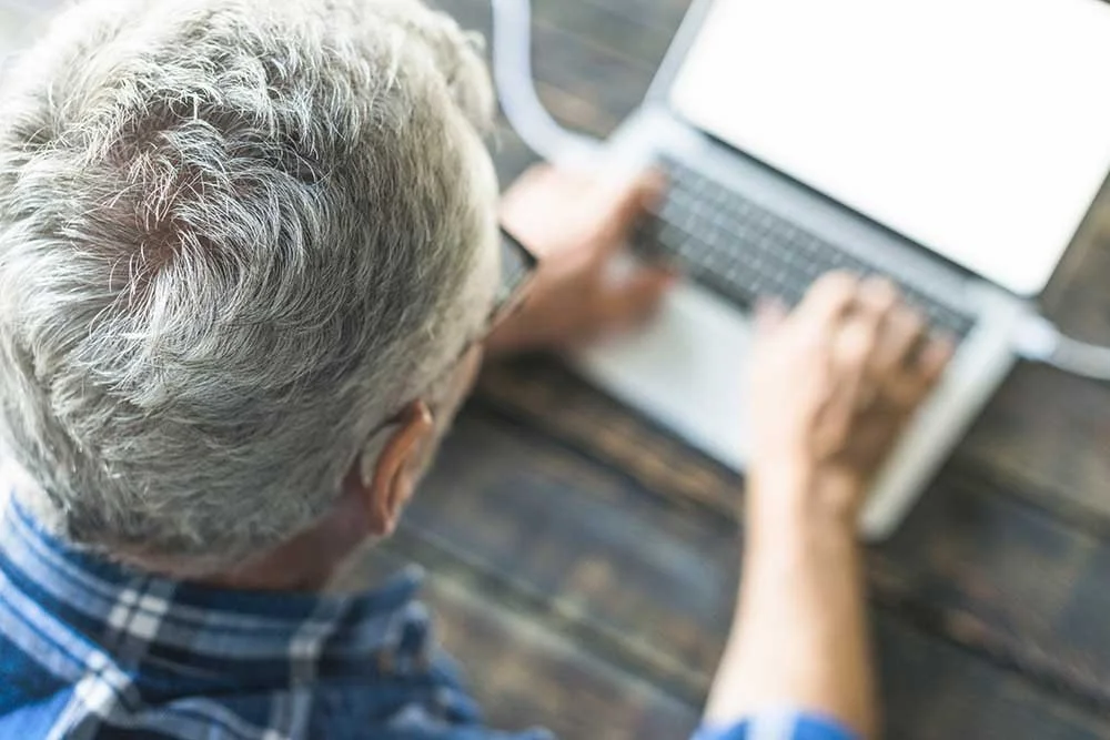 Une personne âgée avec des cheveux gris utilisant un ordinateur portable sur une table en bois, signifiant une aide informatique
