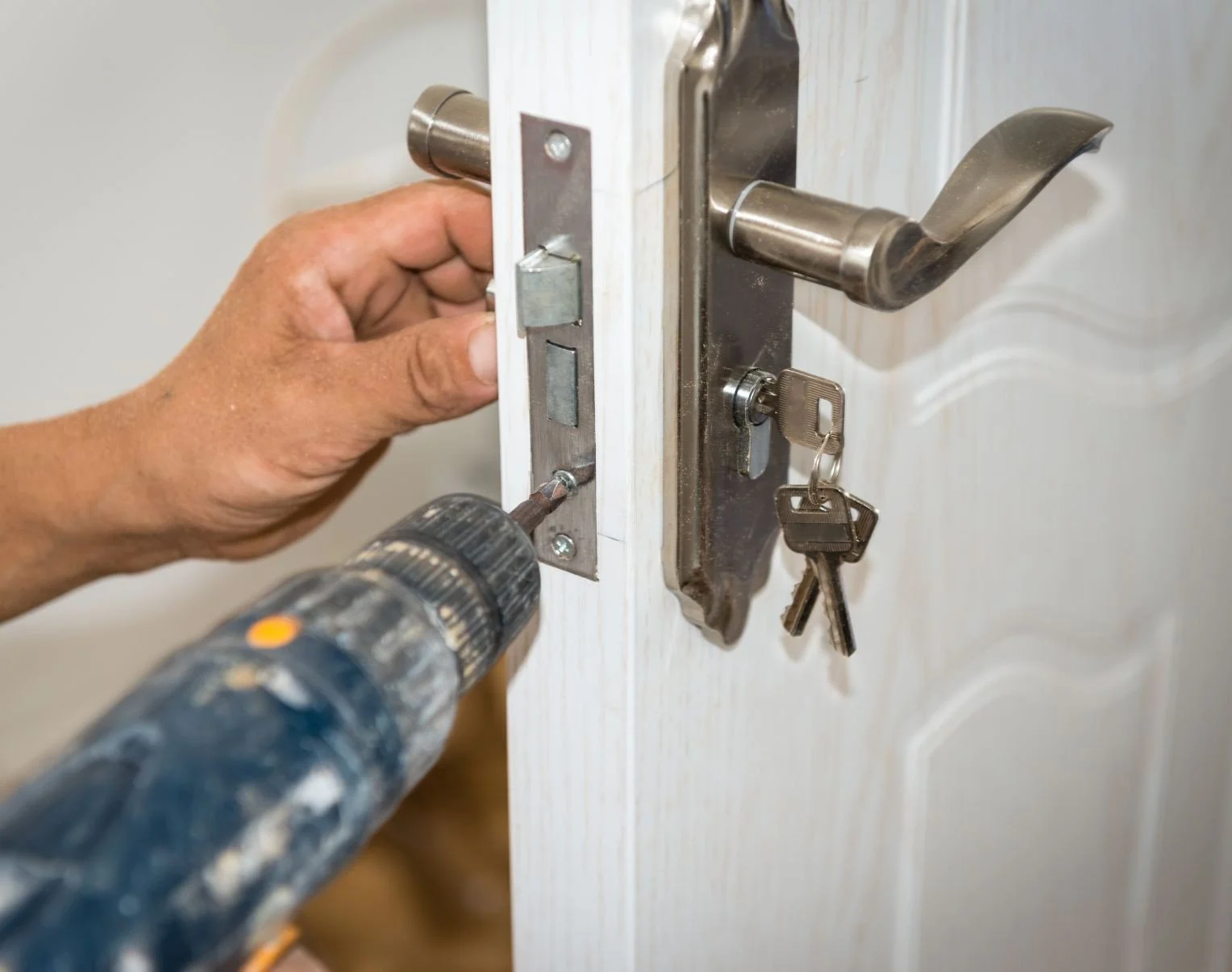 Une main utilise une perceuse pour percer le cylindre du client endommagé sur une porte en bois blanc. Un trousseau de clés est suspendu à la poignée.