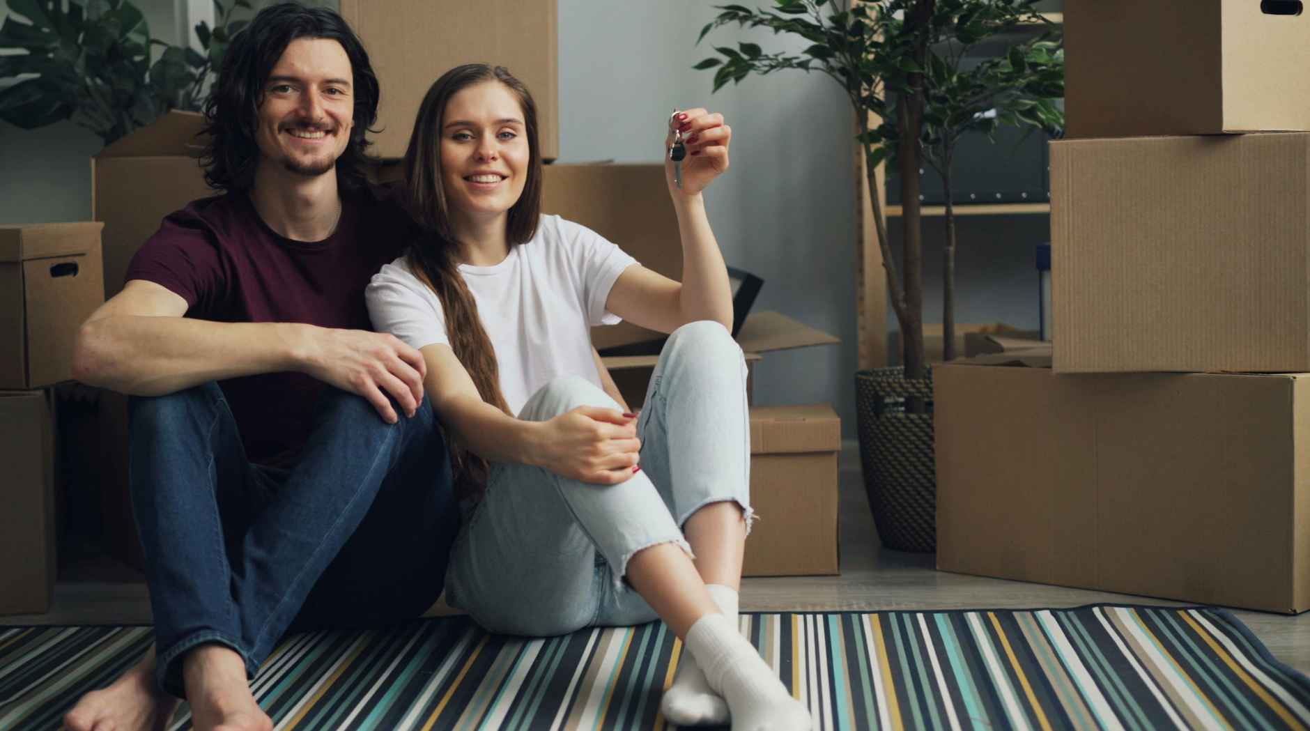 Un jeune couple souriant, assis par terre dans un nouvel appartement, entouré de cartons, avec la femme tenant une clé.