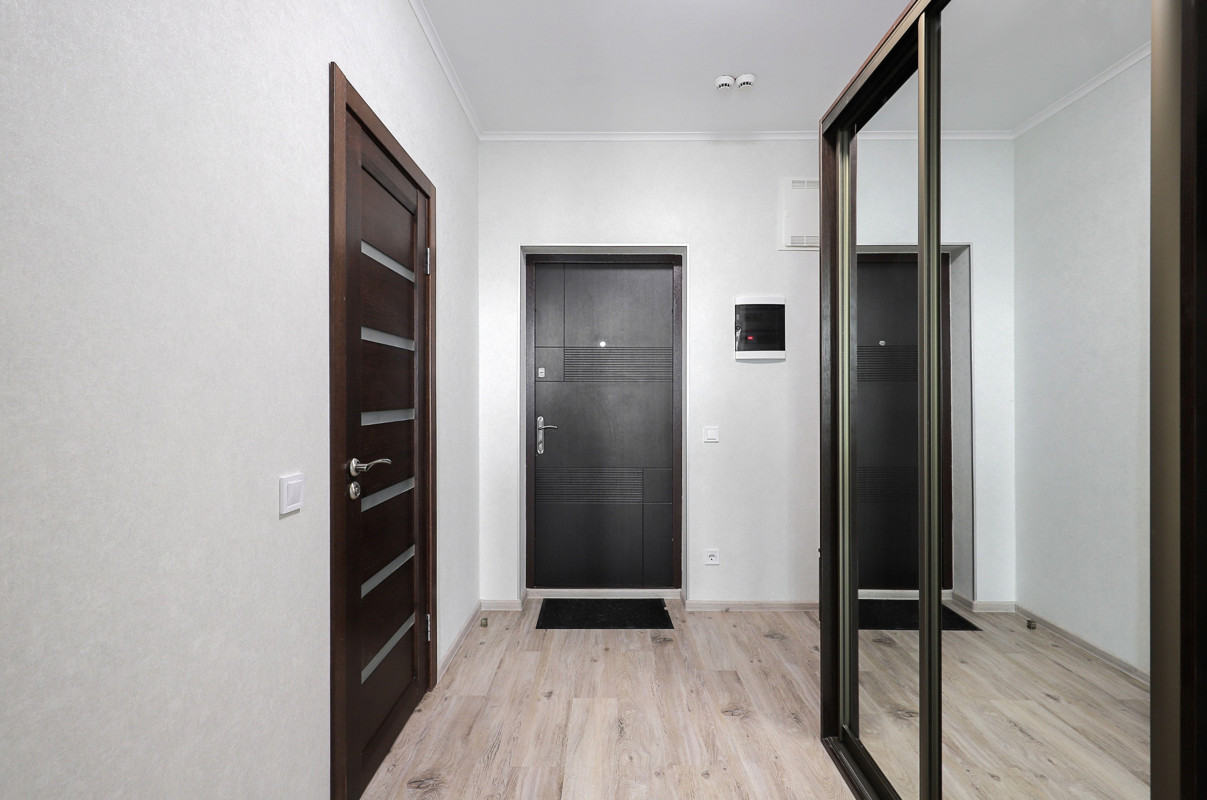 Un hall d'entrée avec un sol en bois clair, deux portes en bois sombre, une porte d'entrée principale en métal noire, un miroir mural, et un meuble à miroir.