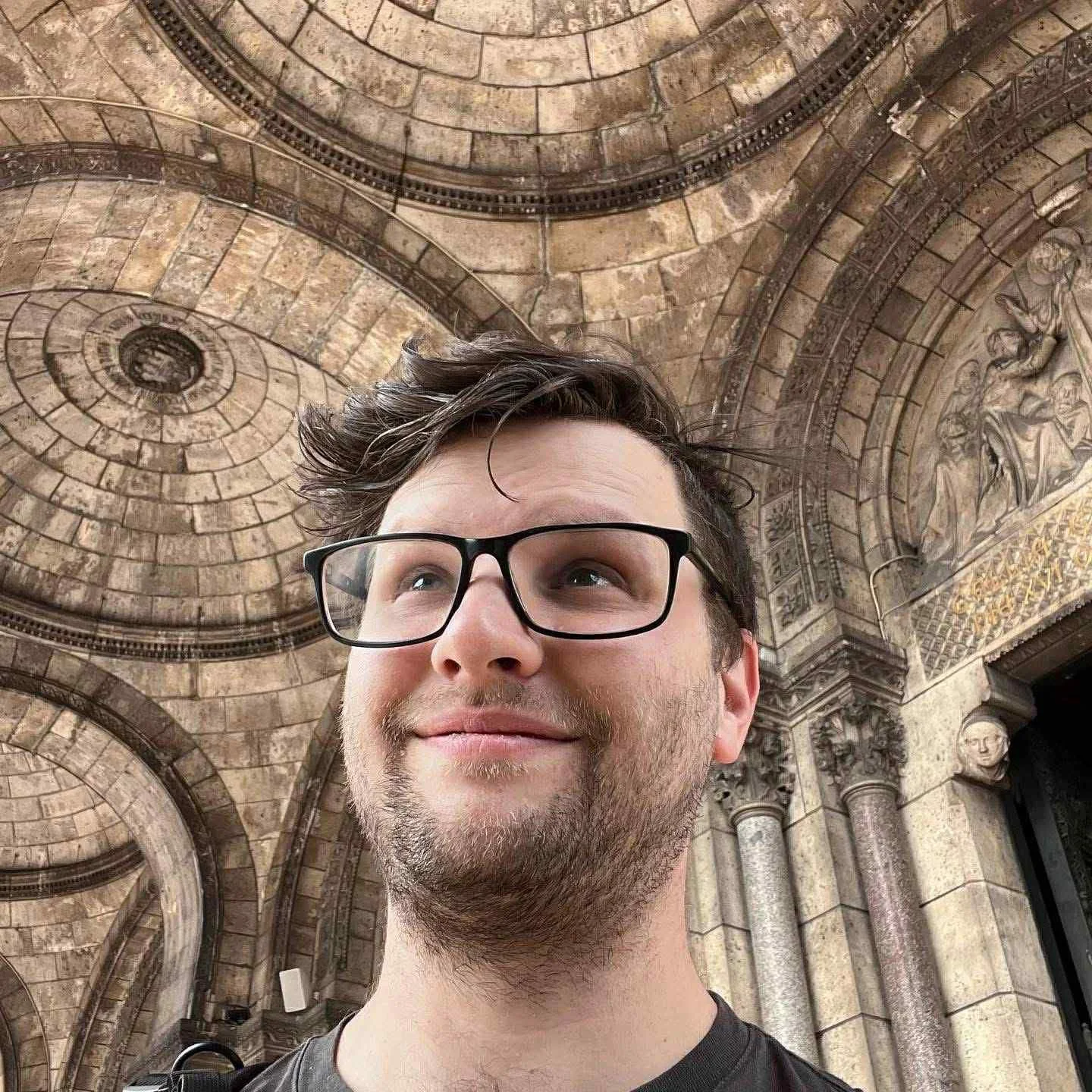 A man with glasses and a beard smiling in front of a historic stone building with ornate architectural details and sculptures.