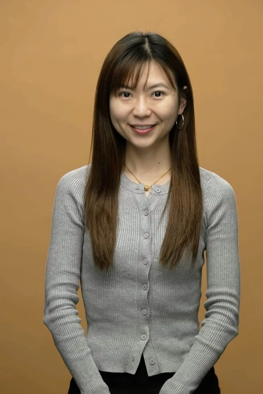 A young woman with long brown hair, wearing a gray button-up cardigan, gold necklace, and hoop earrings, smiling at the camera against a beige background.